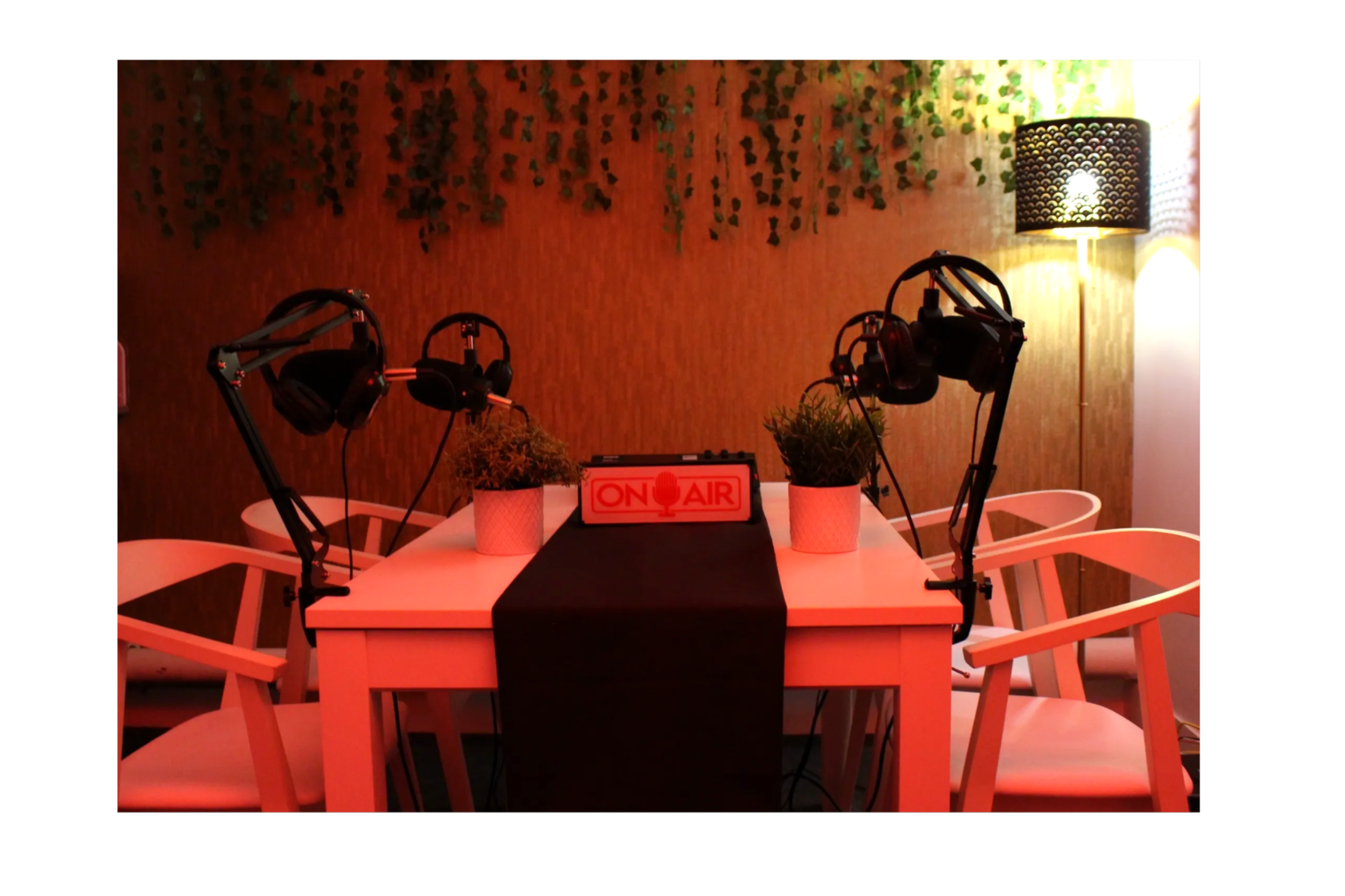 A small podcasting studio features a white table with two microphones and headphones, surrounded by potted plants and illuminated by a lamp with a woven shade.