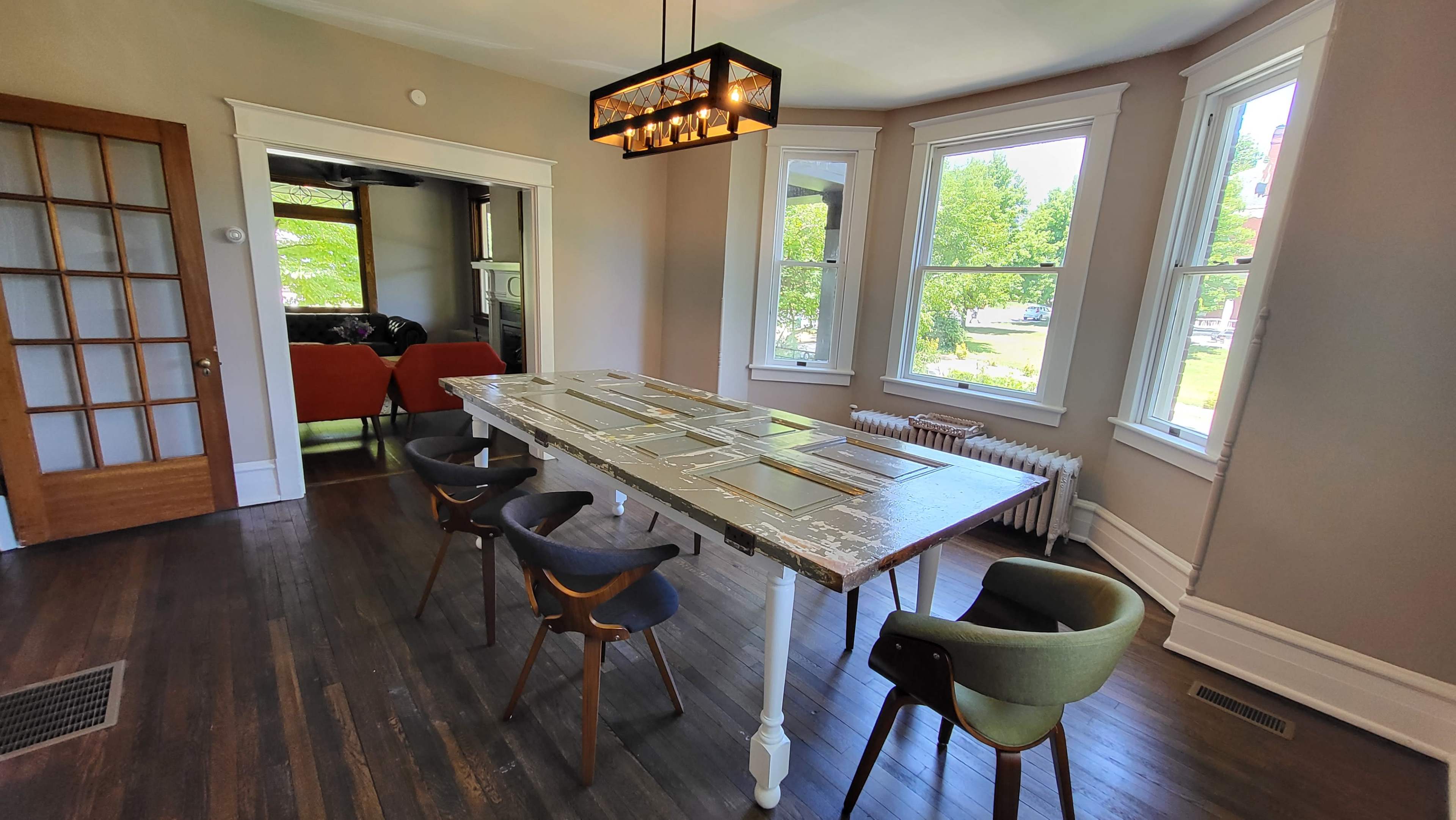 A bright dining room featuring a long, rustic table surrounded by modern chairs, with large windows allowing natural light to enter.