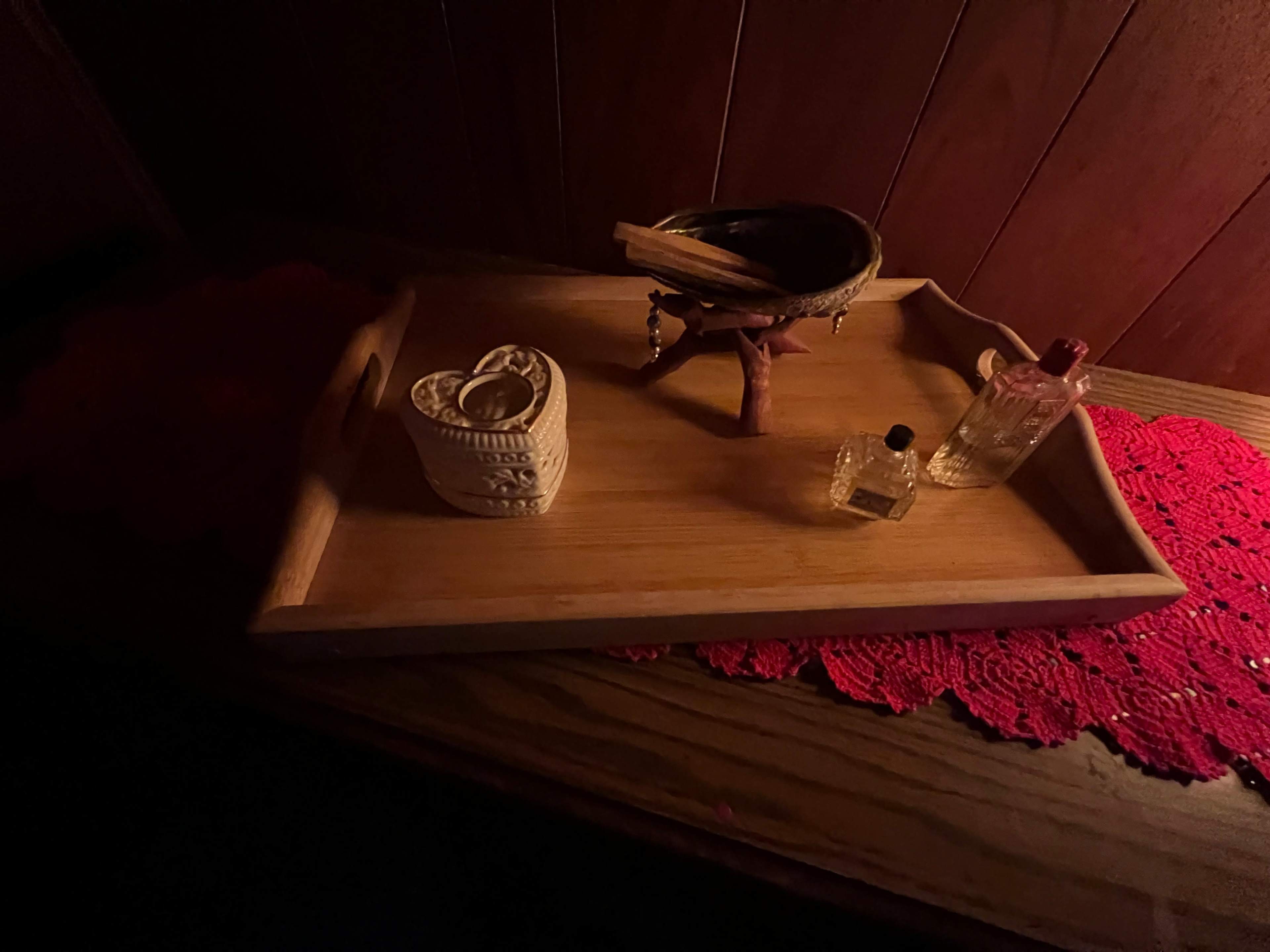 A wooden tray holds a decorative bowl, two small glass bottles, and a carved heart-shaped item, all set on a red lace doily.