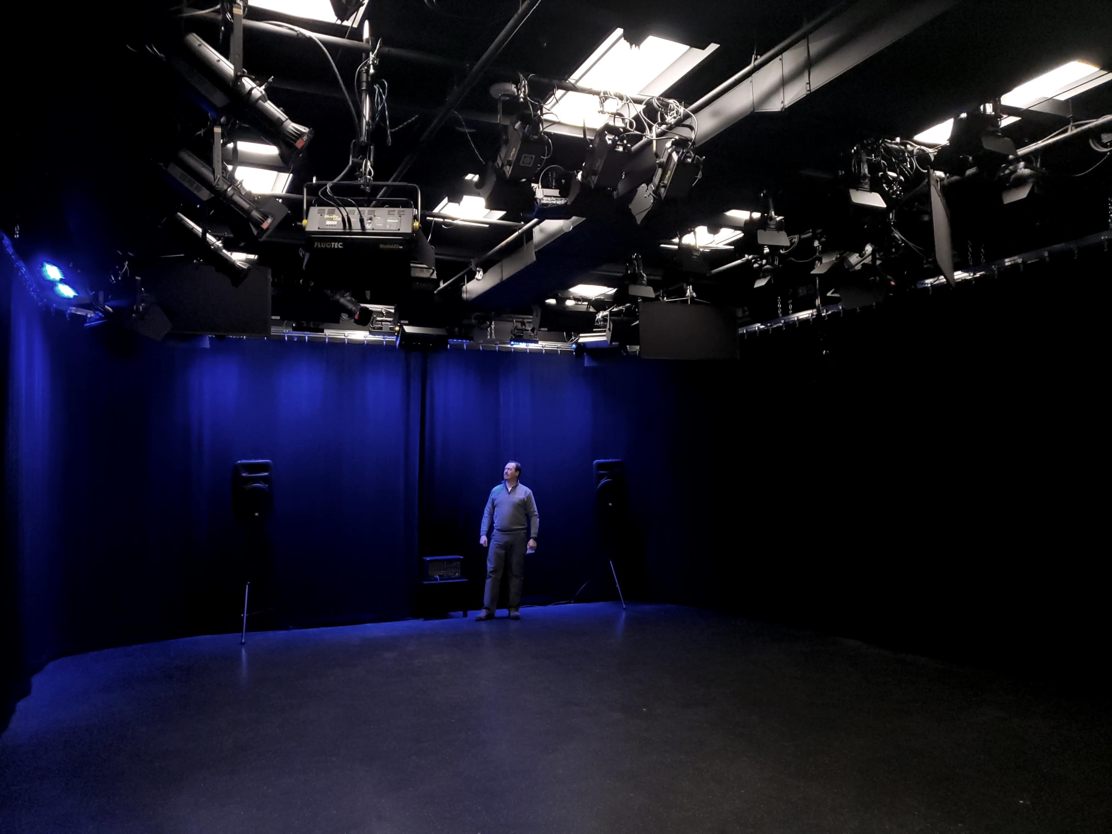 A man stands alone in a dimly lit studio with black curtains and overhead lighting.
