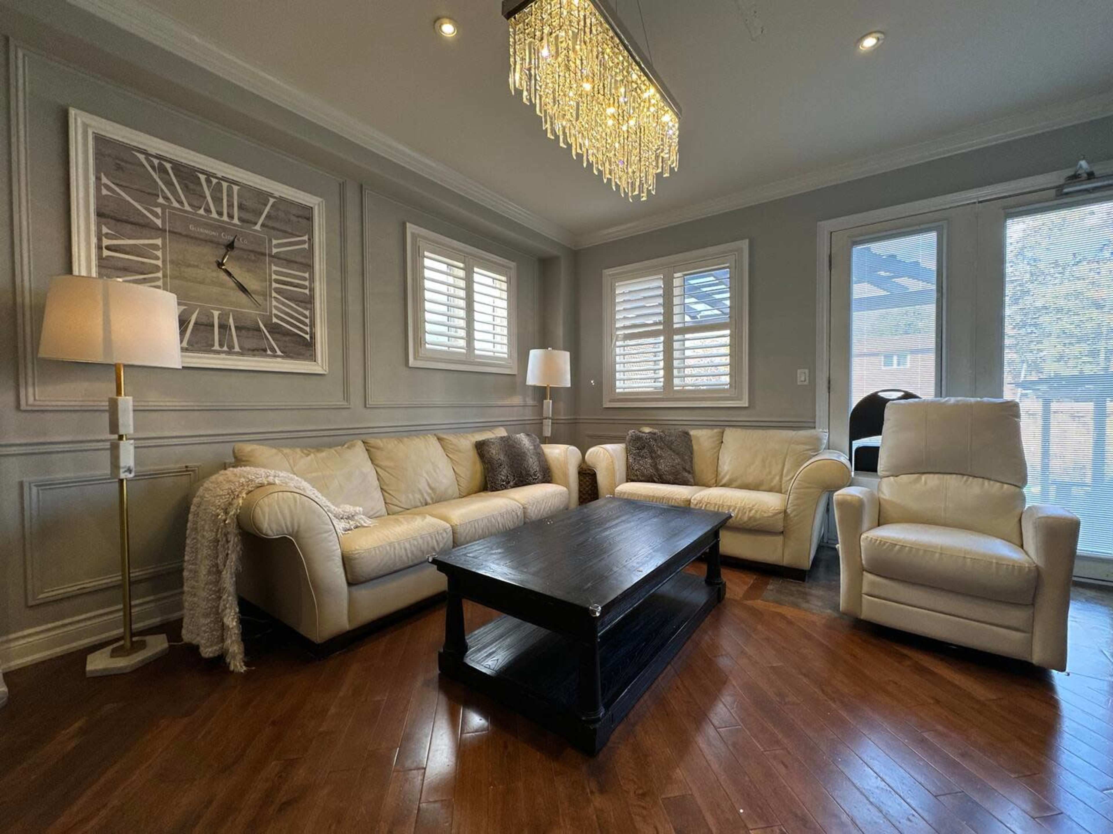 The living room features a white leather sofa set arranged around a dark wooden coffee table, accented by a chandelier and large wall clock.