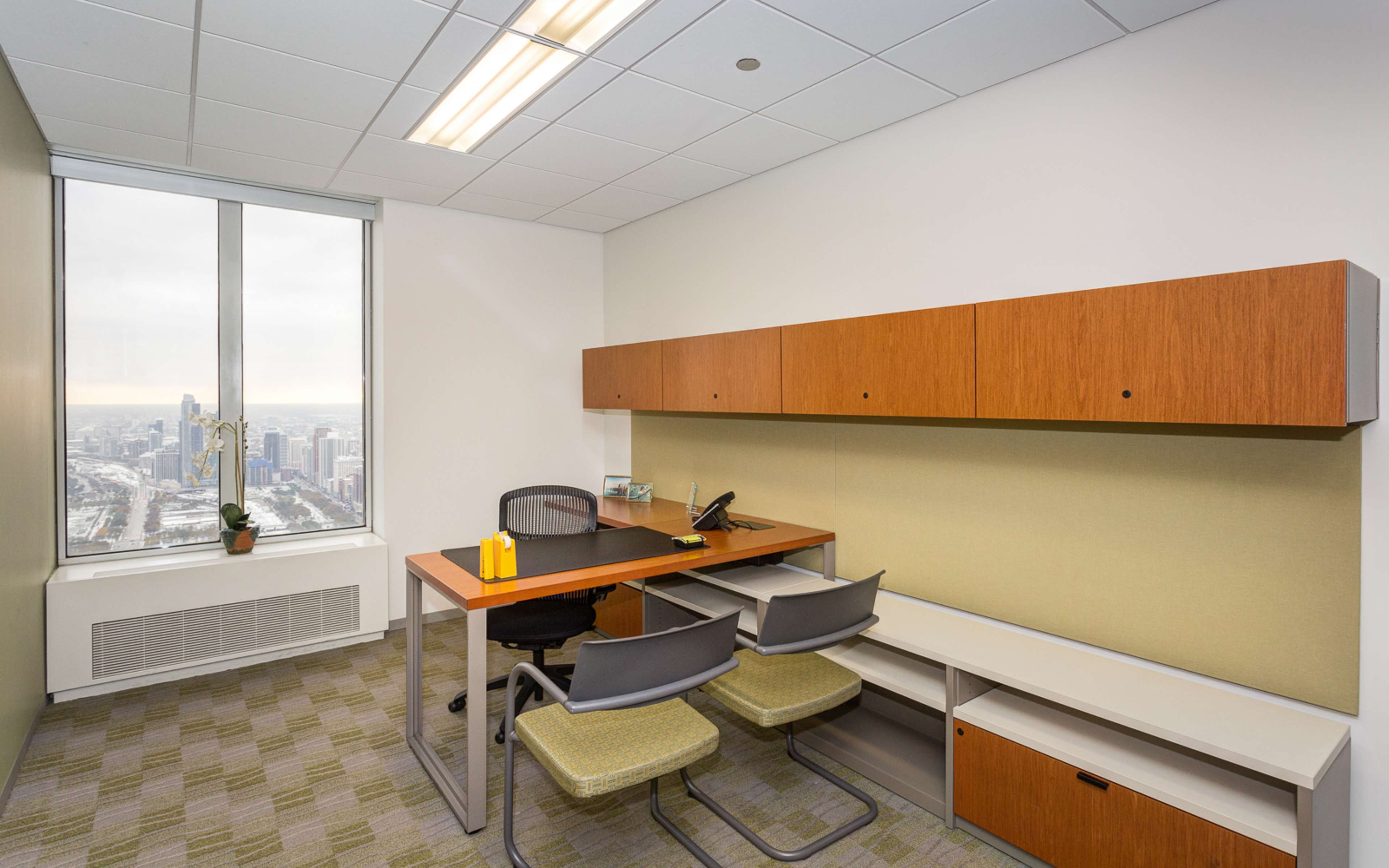 The image shows a modern office space with a desk, two chairs, and storage cabinets, featuring a large window overlooking a city skyline.