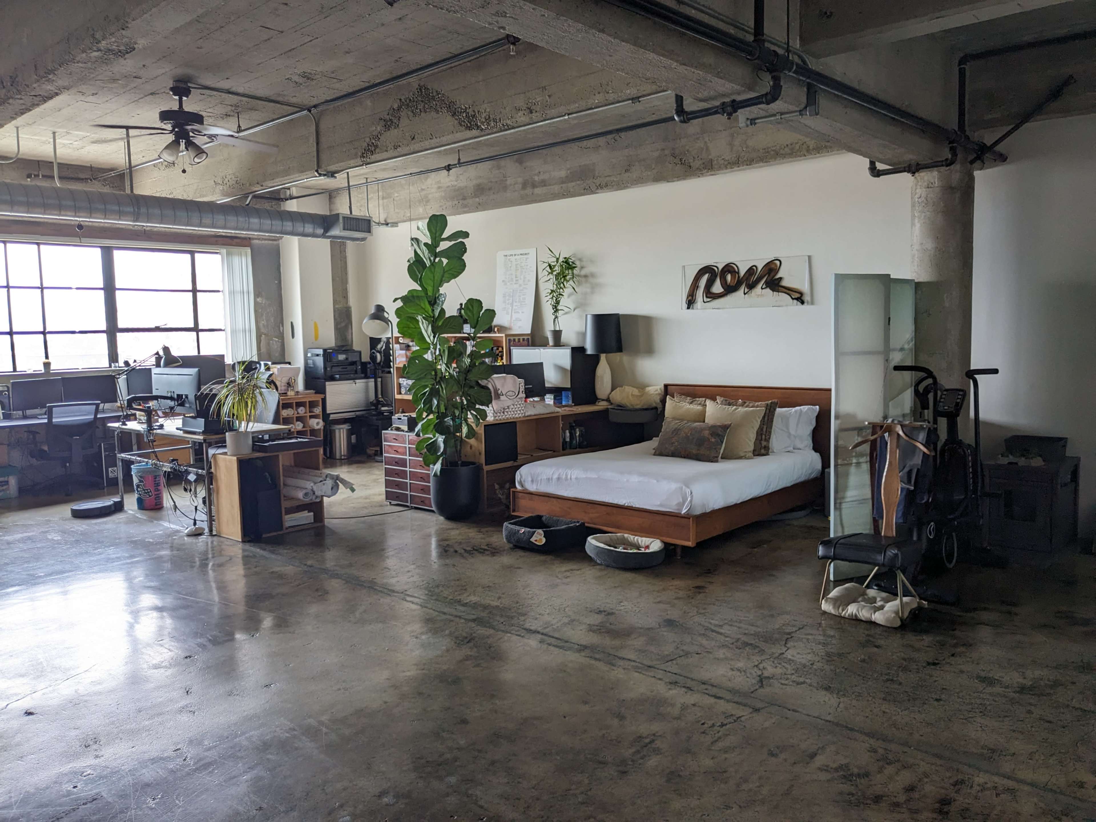 An open loft-style apartment features a bedroom area with a bed, a large window, a desk with computers, and potted plants.