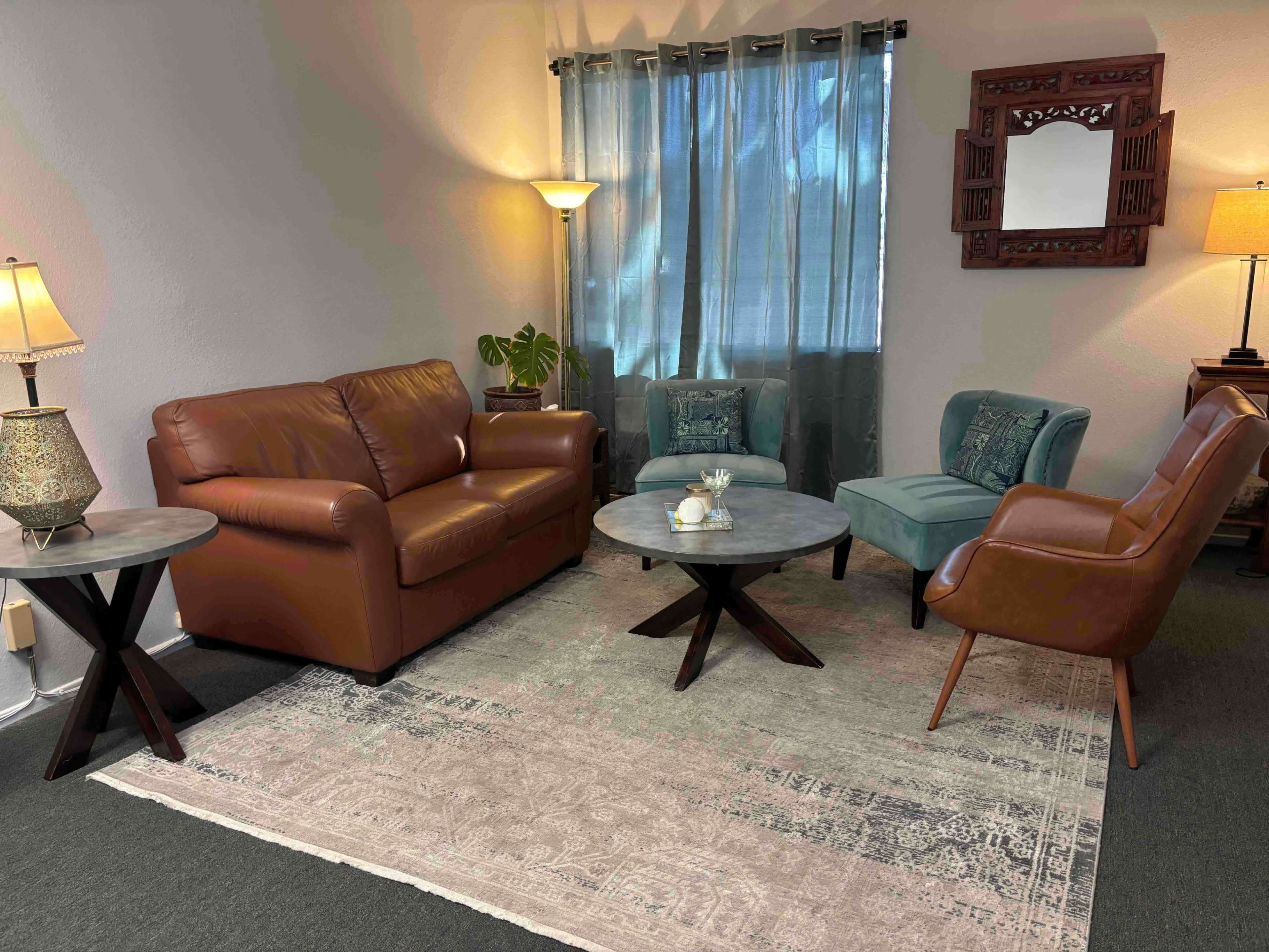 The image shows a cozy living room with a brown leather sofa, two blue upholstered chairs, a round coffee table, and decorative lamps, all arranged on a patterned area rug.