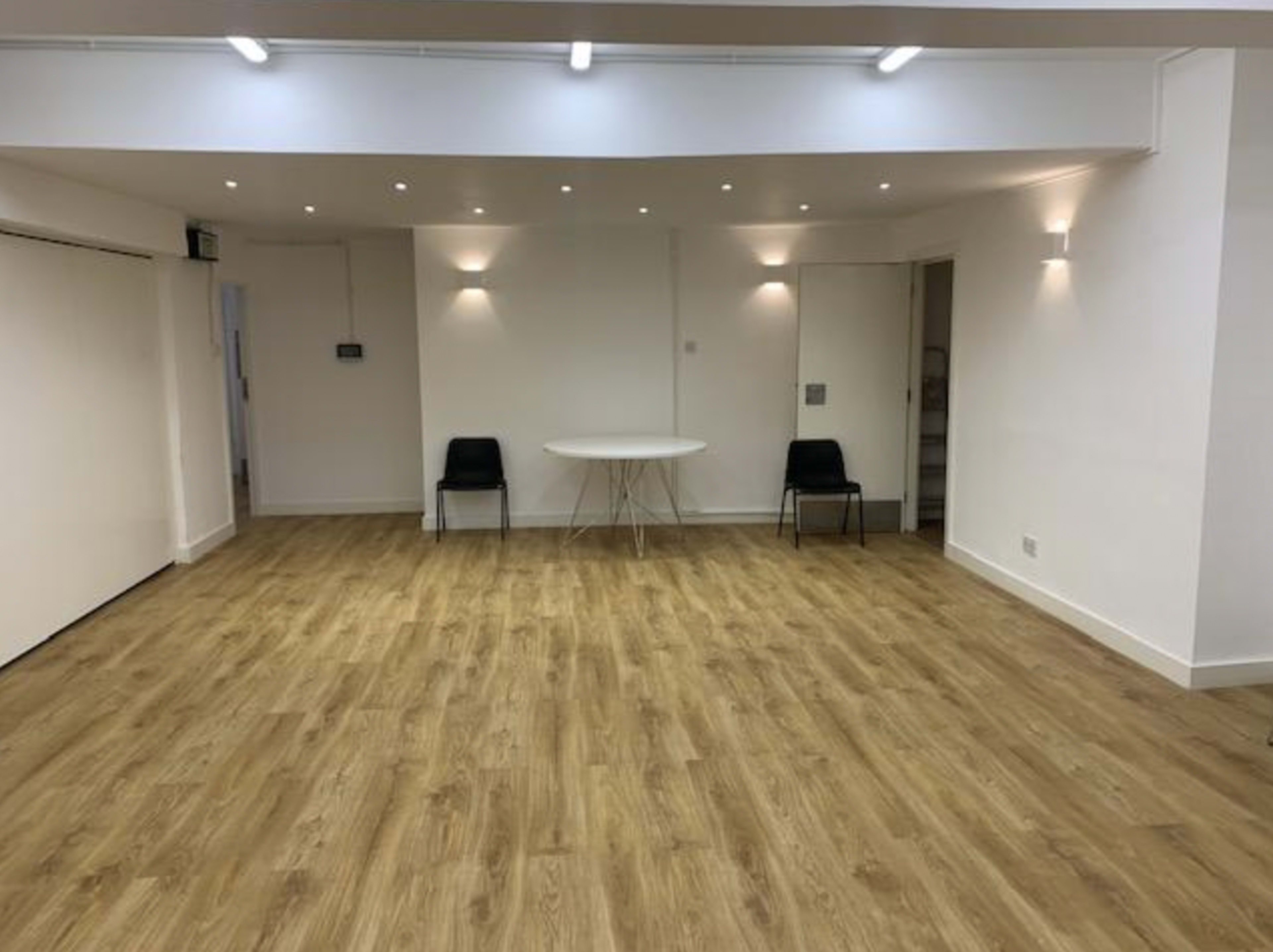 The image shows a spacious, minimalistic room with wooden flooring, featuring a round table in the center and two black chairs positioned nearby.