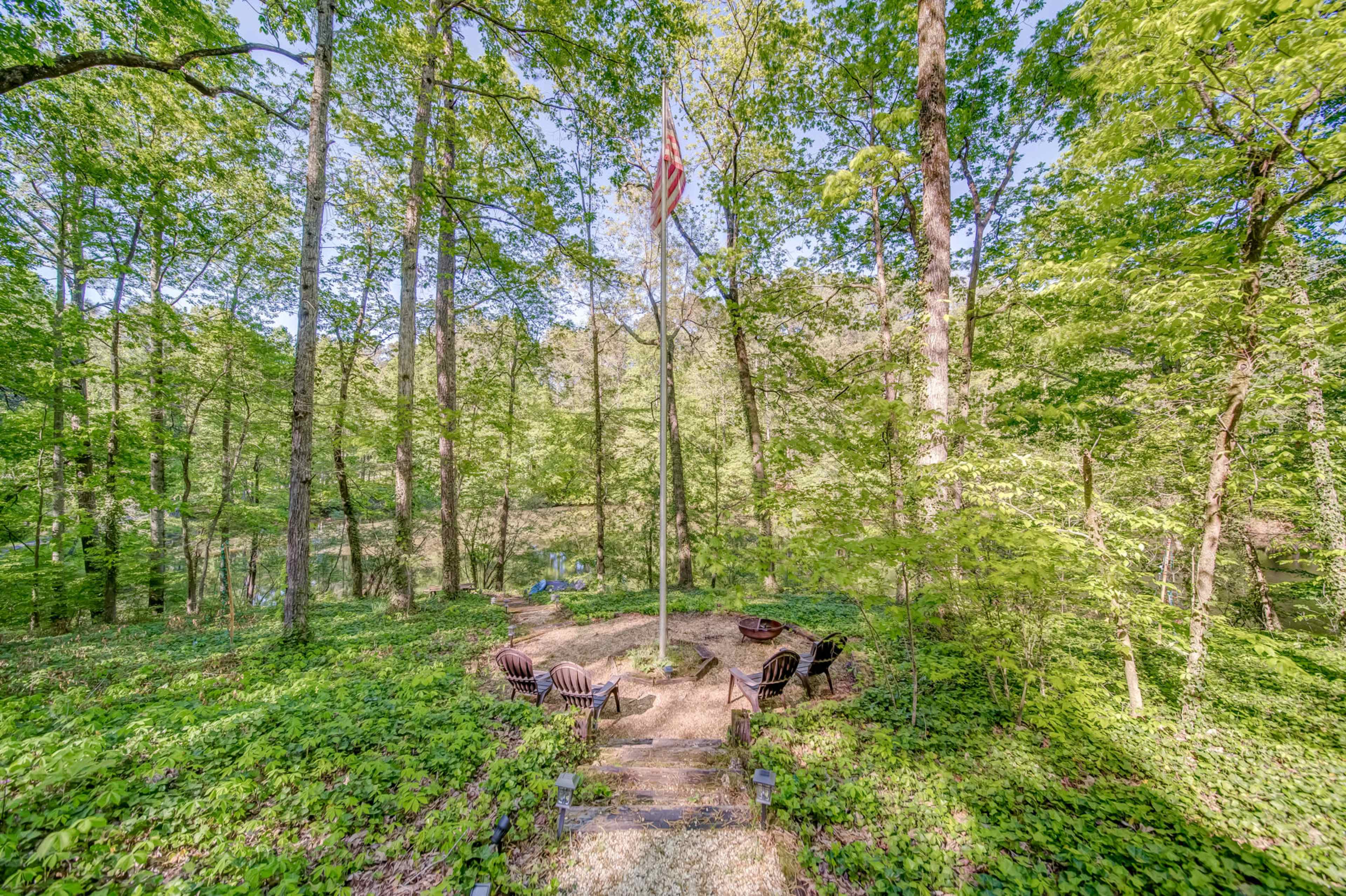 A circular fire pit area with wooden chairs is surrounded by dense green trees in a forested setting.