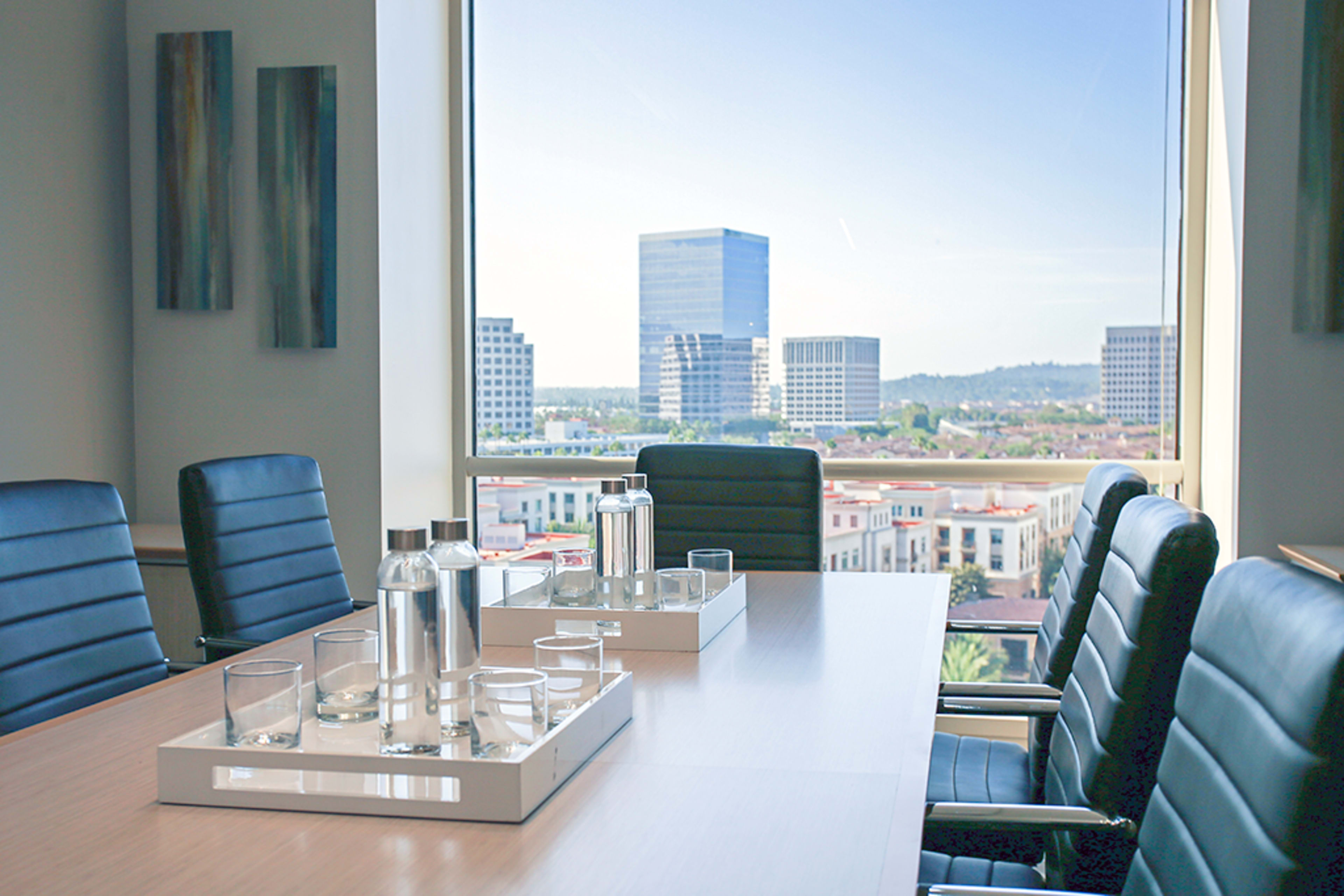 A conference room with a long table, chairs, and a tray of glassware, features large windows overlooking a city skyline.