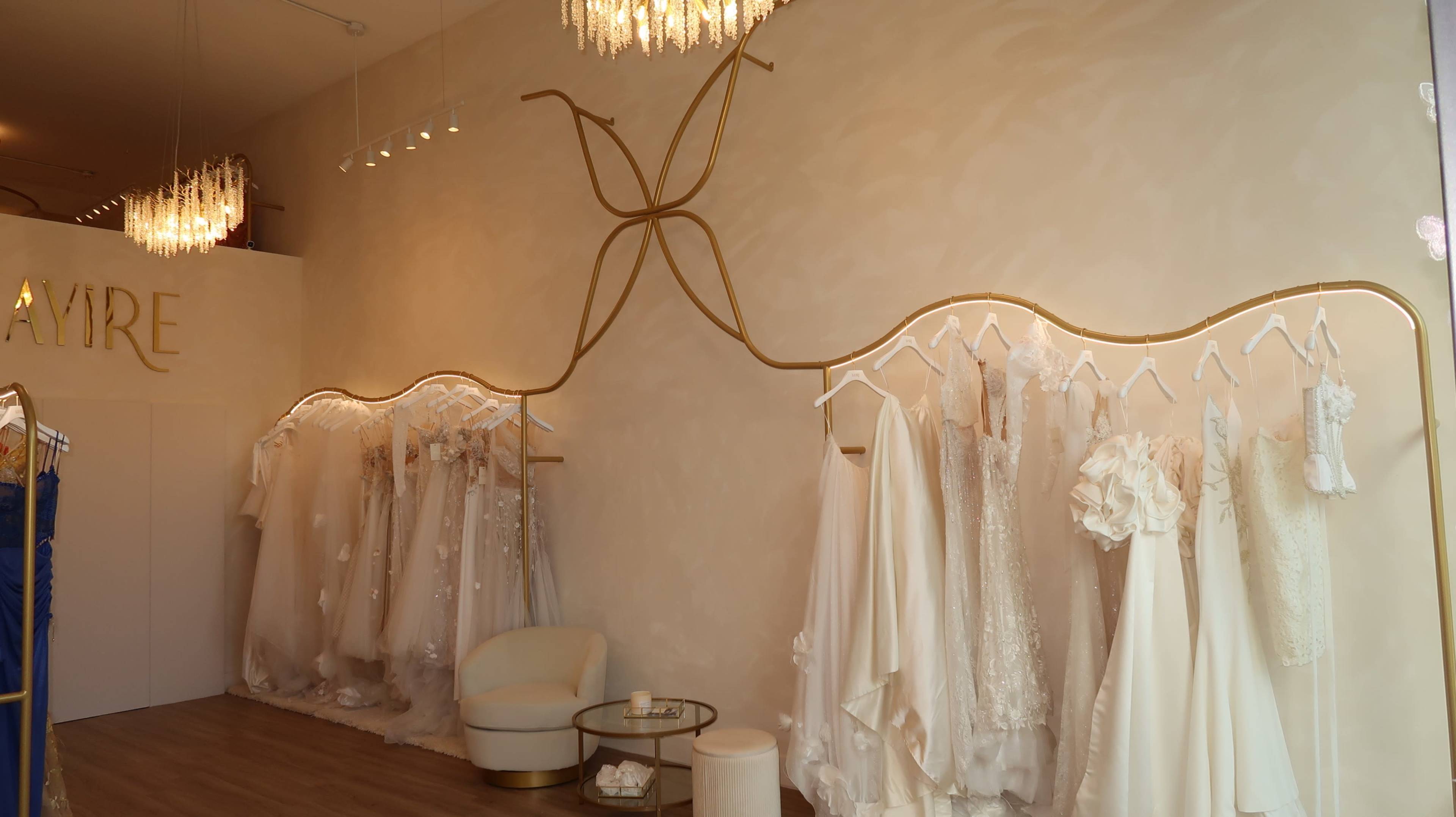 The image shows a bridal shop interior featuring a collection of wedding dresses displayed on hangers along a wall, with elegant lighting and a seating area.