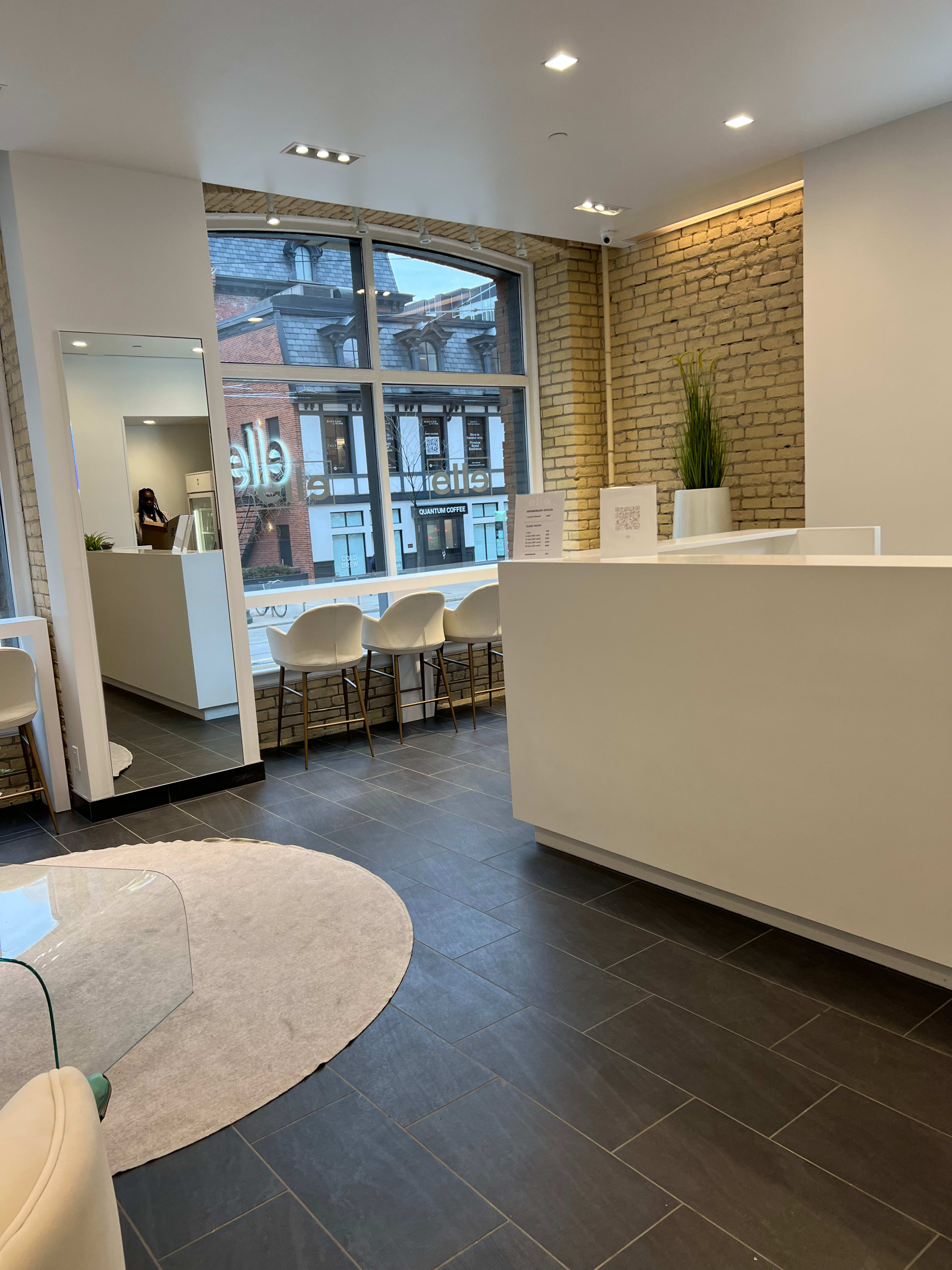A modern reception area with a minimalist design, featuring a white front desk, seating arranged along a window, and a large mirror reflecting the space.