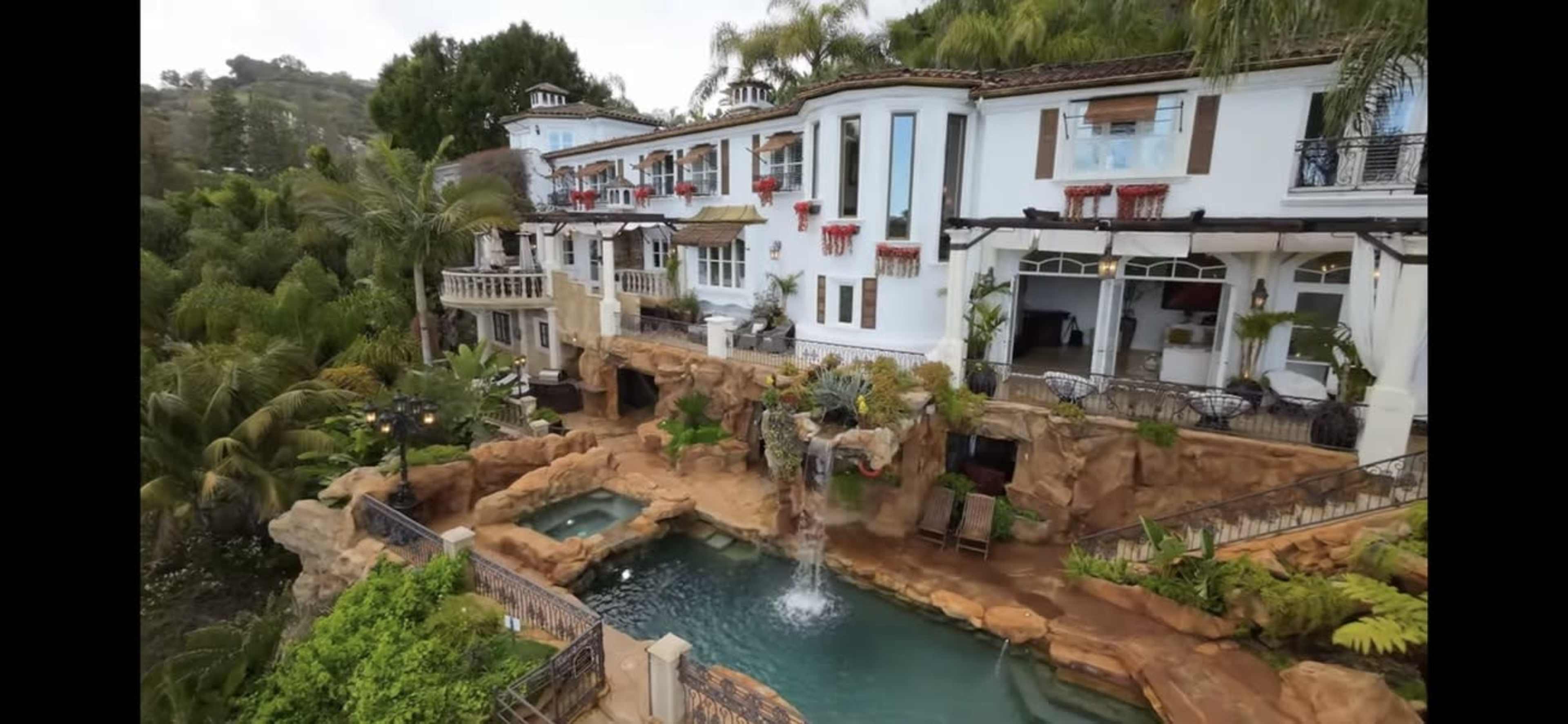 A luxury house with multiple levels, surrounded by palm trees and a rocky waterfall that flows into a swimming pool.