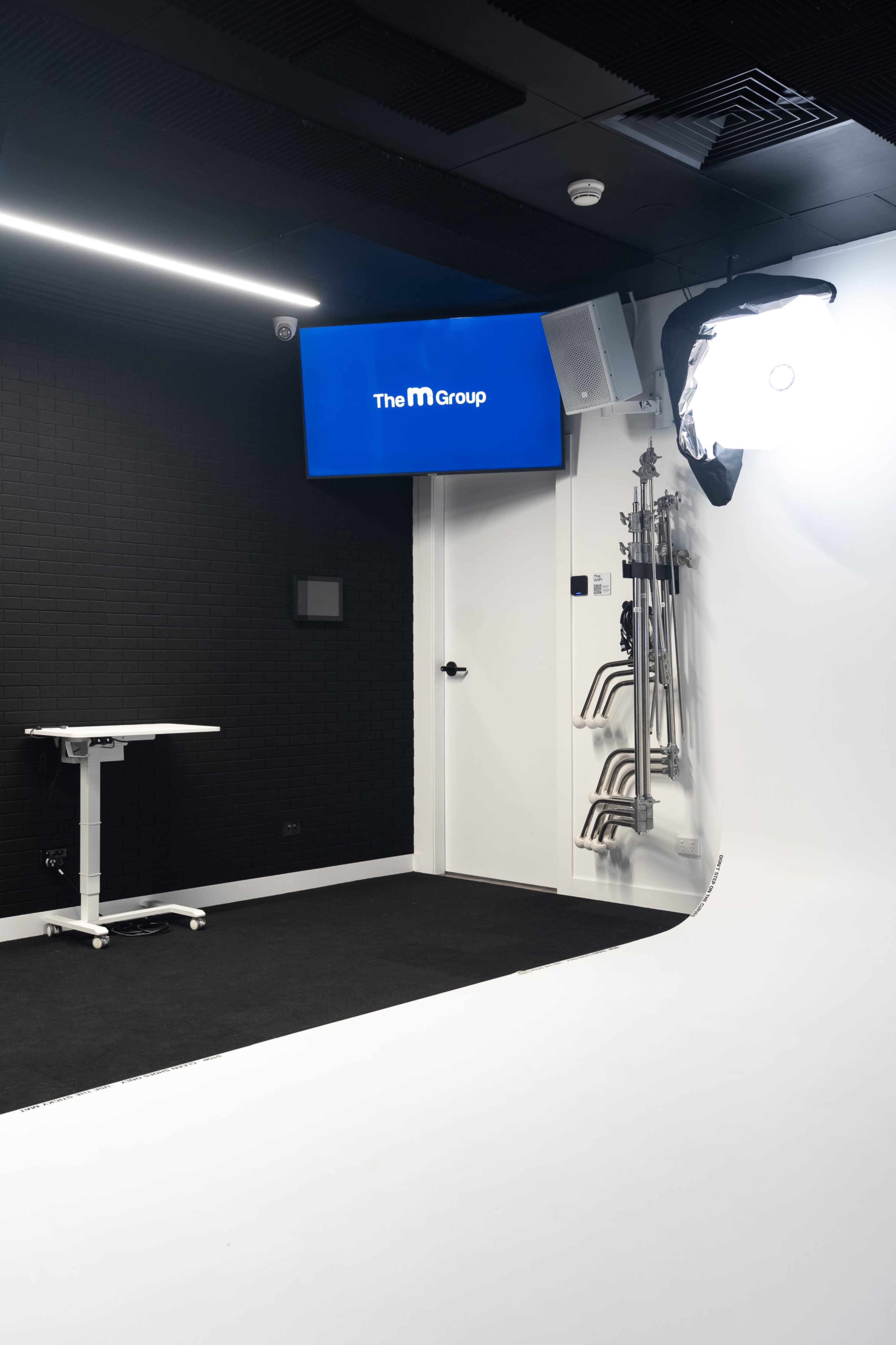 The image shows a modern studio setup featuring a large screen displaying "The M Group," a movable table, and several microphone stands against a black wall.