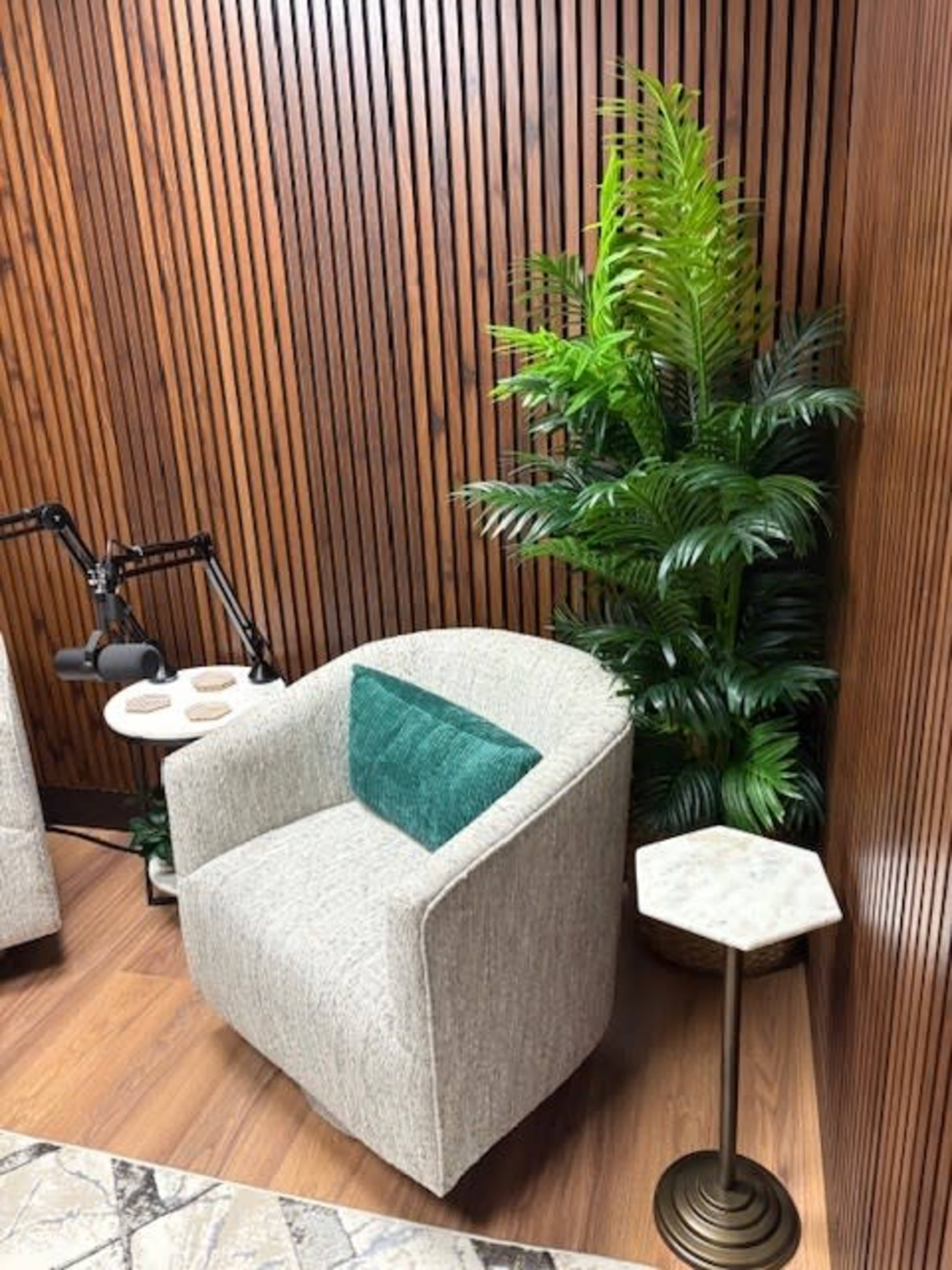 The image shows a cozy corner with a light-colored armchair featuring a teal cushion, a small marble-topped side table, a modern black lamp, and a lush green plant against a wooden panel wall.