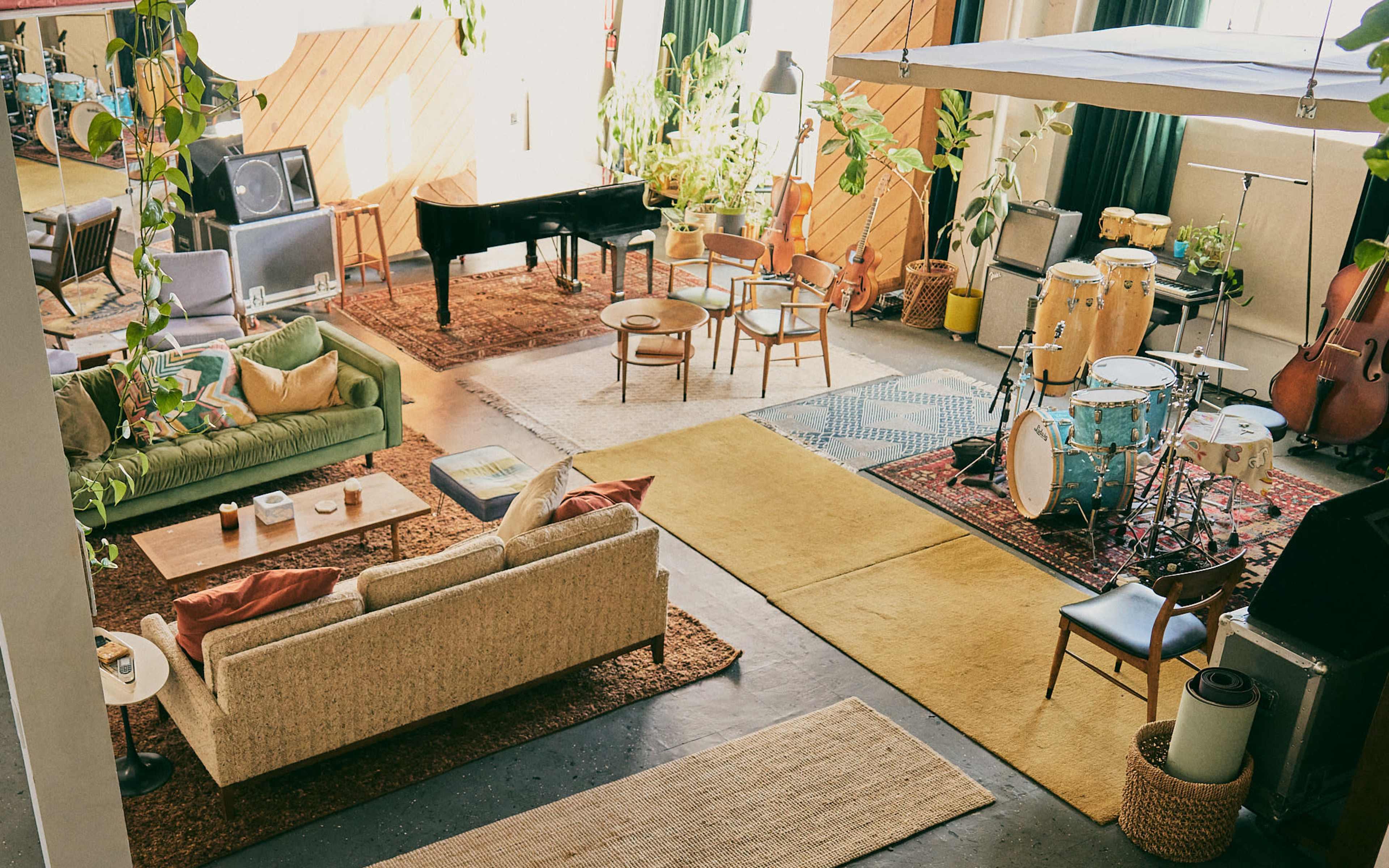 The image shows a bright, spacious room with various seating areas, musical instruments, and plants throughout.