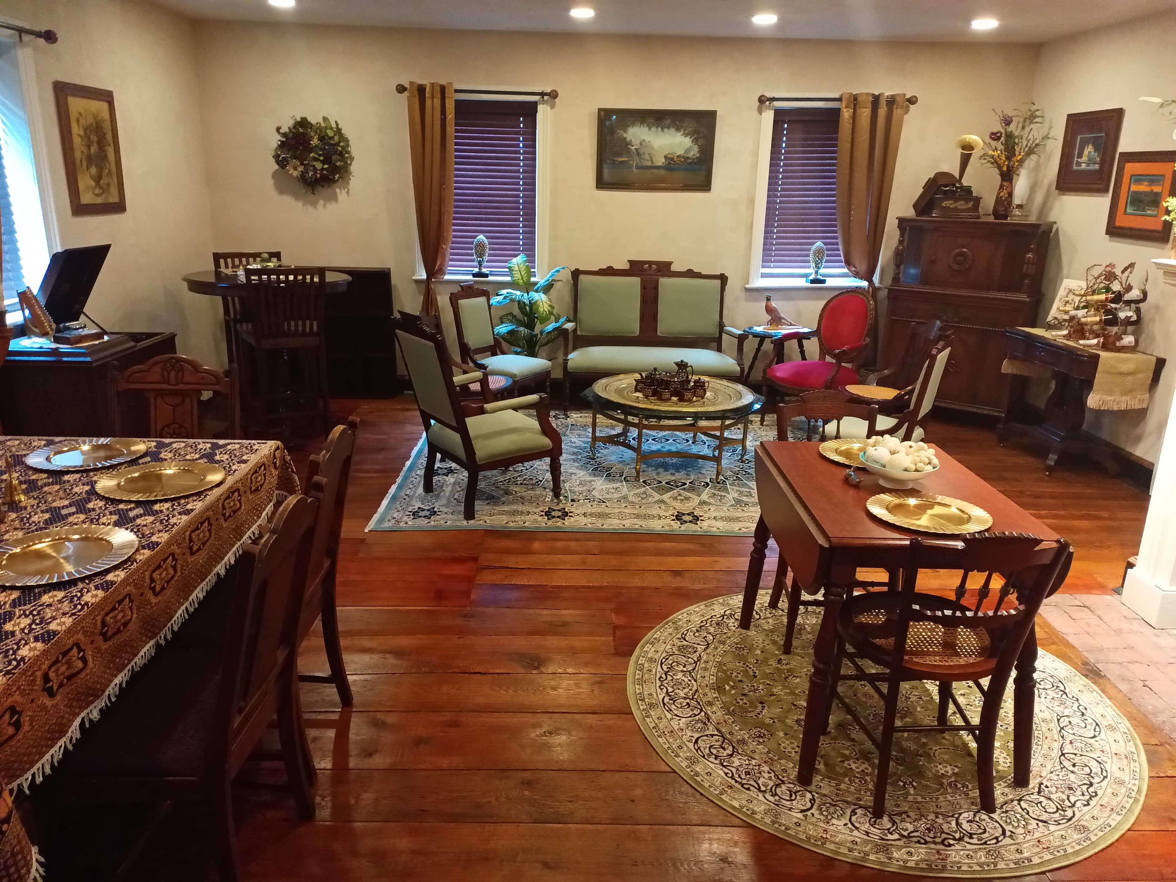 A vintage-style living room and dining area with wooden floors, a dining table set for a meal, and various antique furniture pieces arranged around the space.