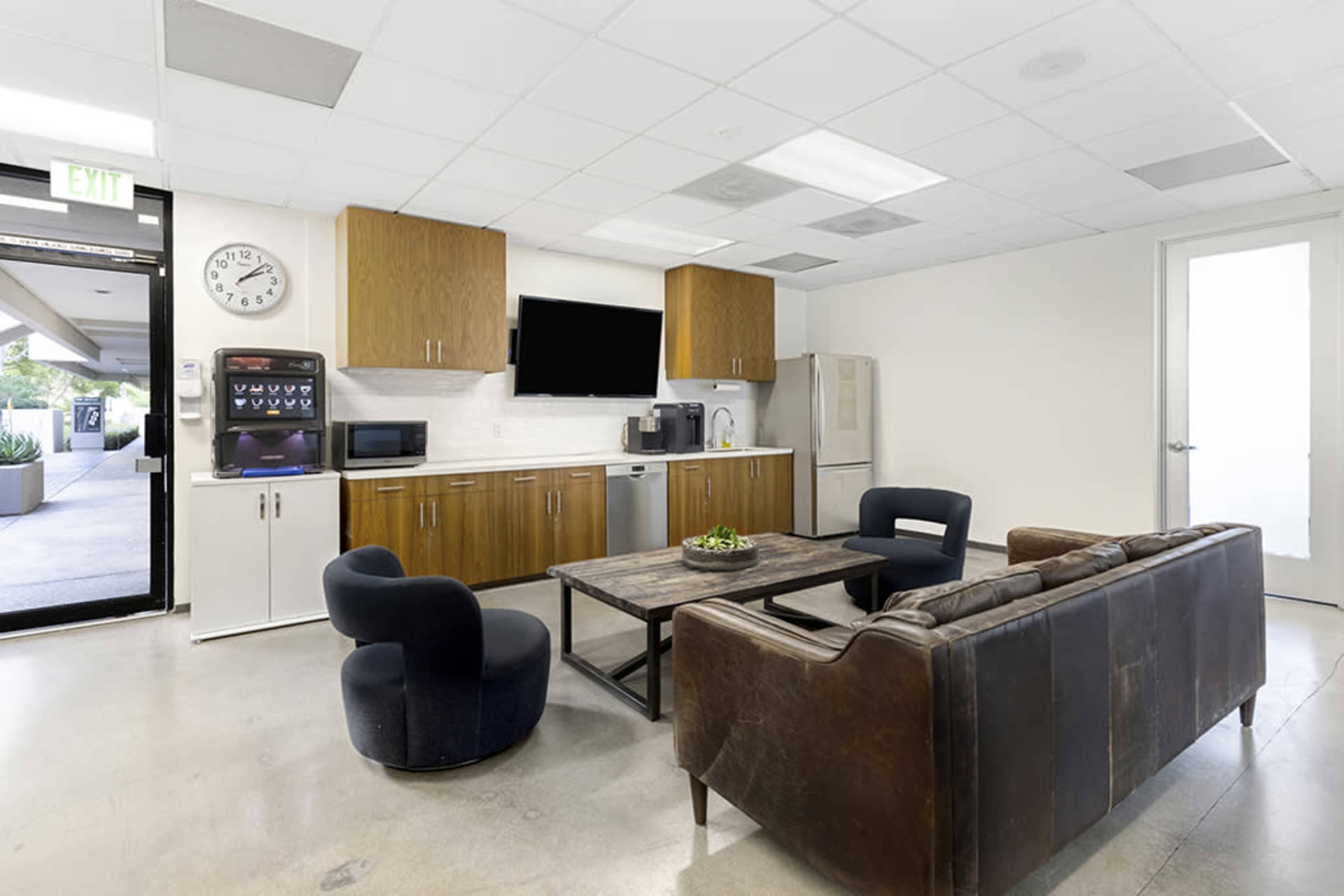 A modern break room with a couch, chairs, table, kitchen appliances, and a wall-mounted television.