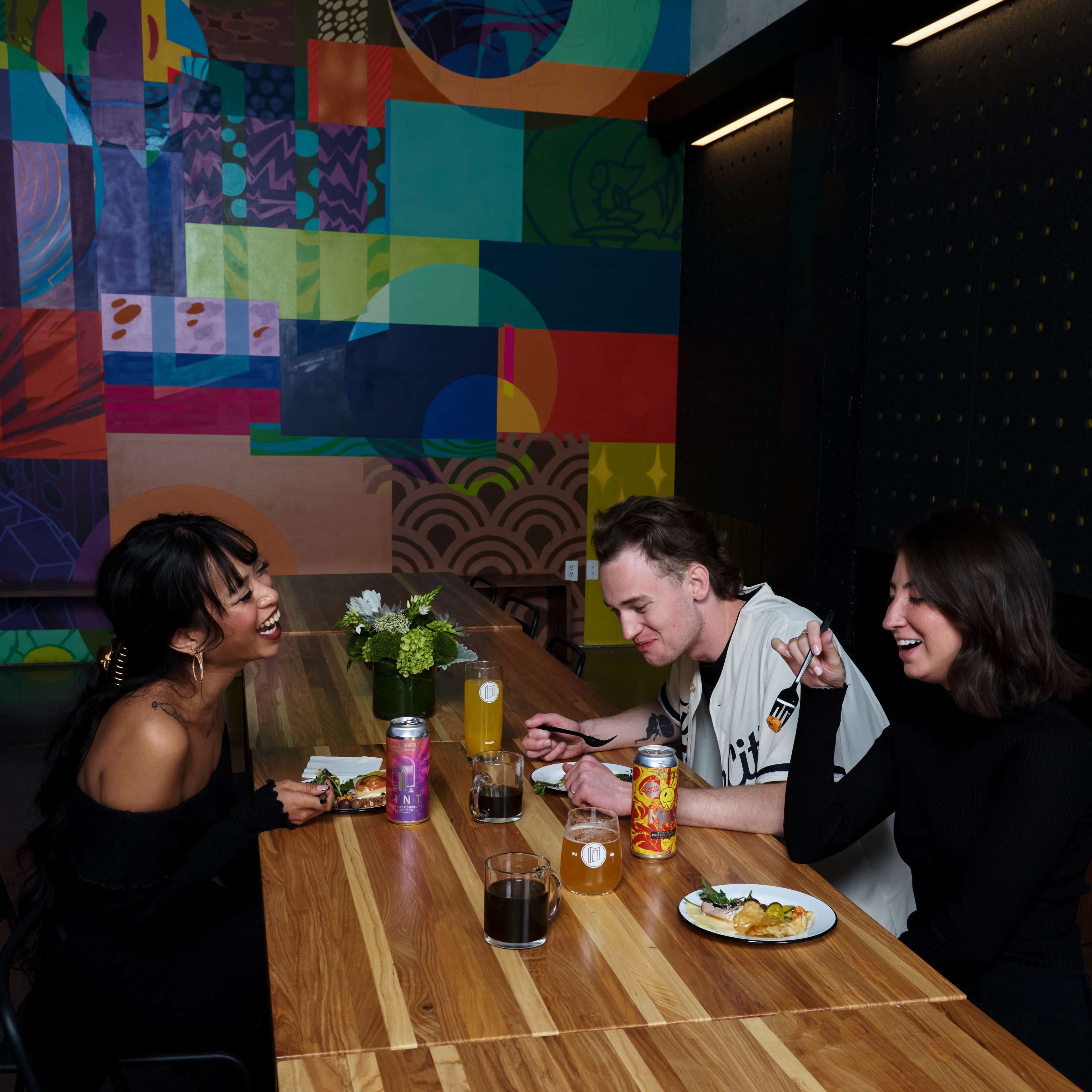 Three people are seated around a wooden table, enjoying food and drinks in front of a colorful mural.
