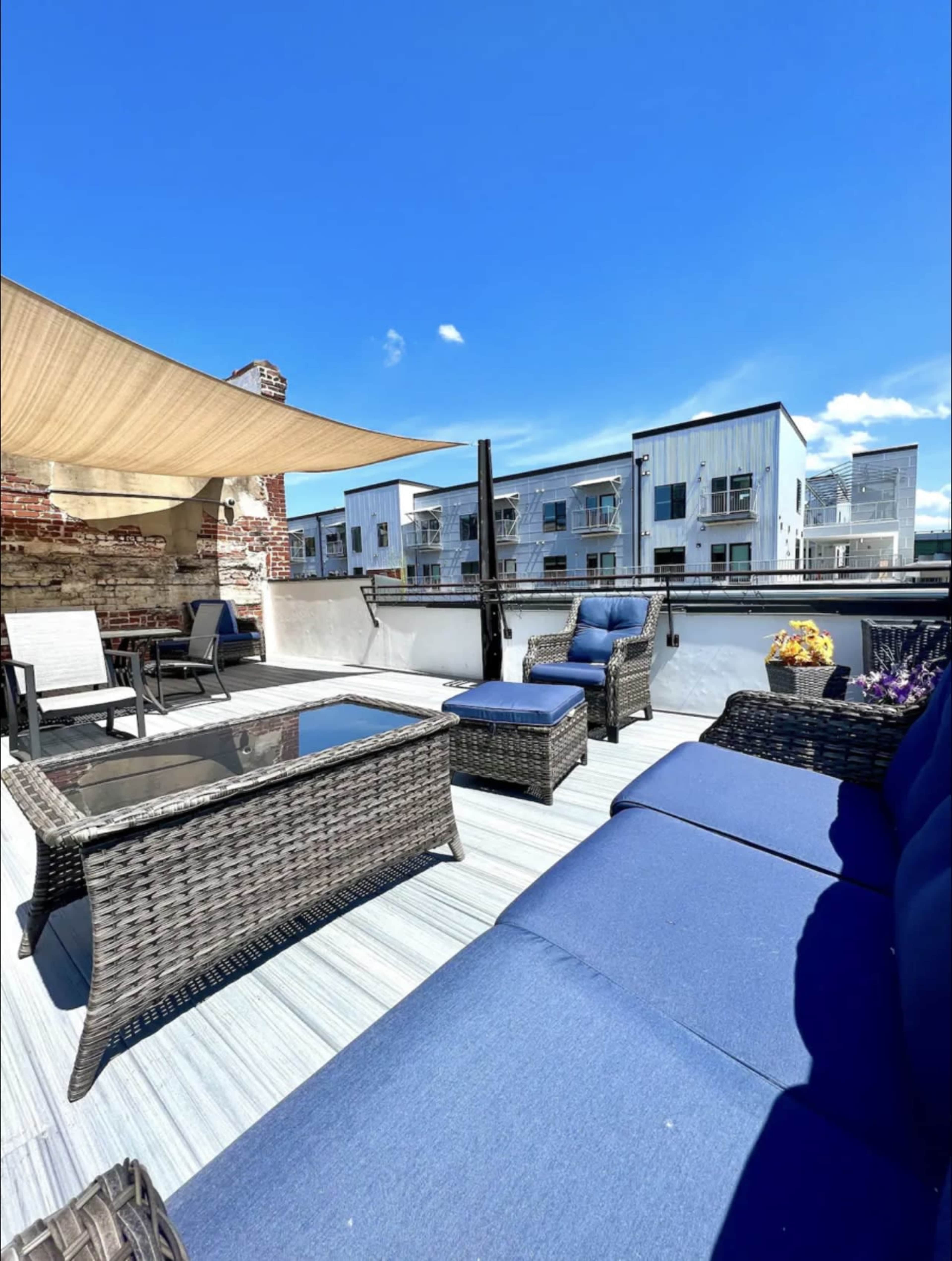 A rooftop patio with wicker furniture, a shaded area, and a view of nearby modern buildings under a clear blue sky.
