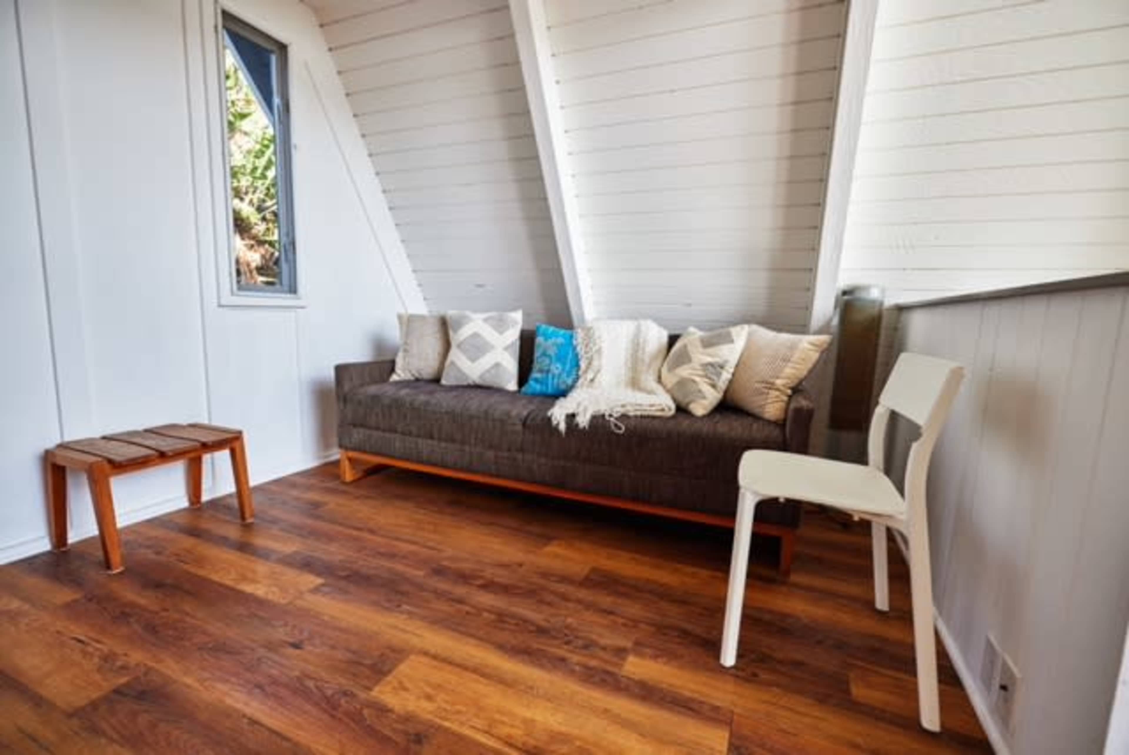 The image shows a small, modern living space featuring a gray couch with decorative pillows, a wooden side table, and a white chair against a slanted wall.