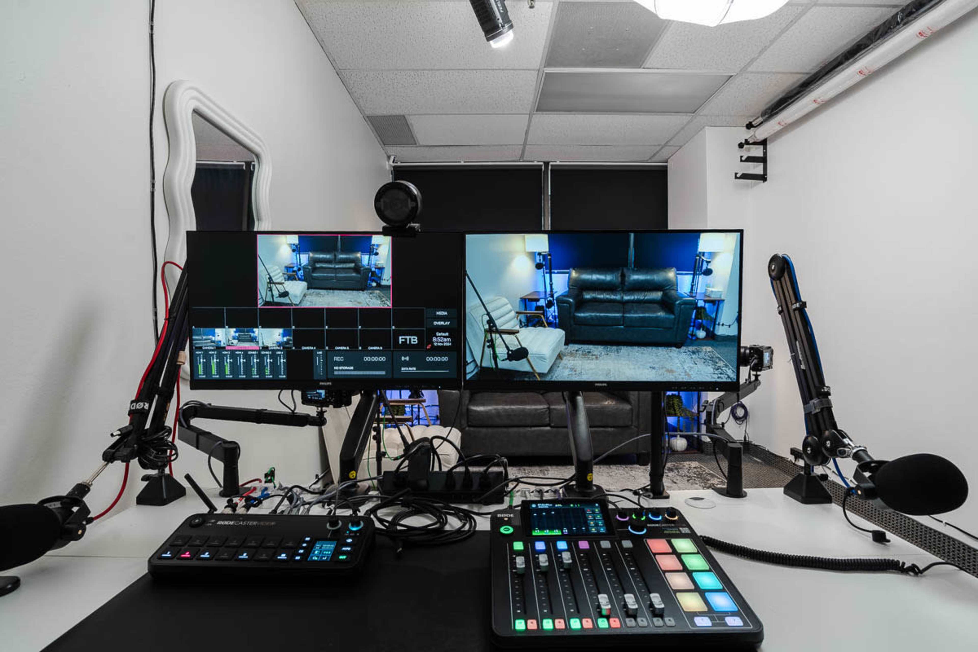 A broadcasting studio setup features two monitors displaying a live feed of a room with a couch, alongside audio equipment and control panels on a desk.