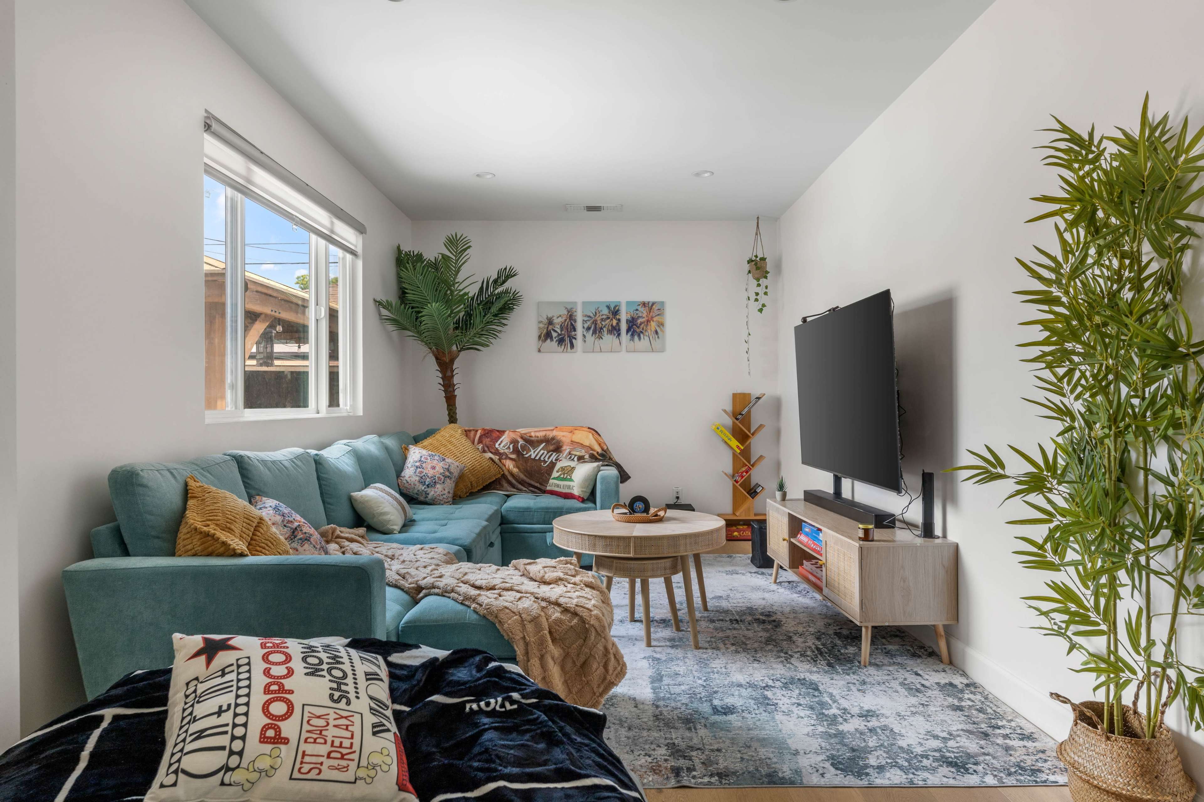 The image shows a modern living room with a large sectional sofa, a coffee table, and a flat-screen TV, surrounded by decorative plants.