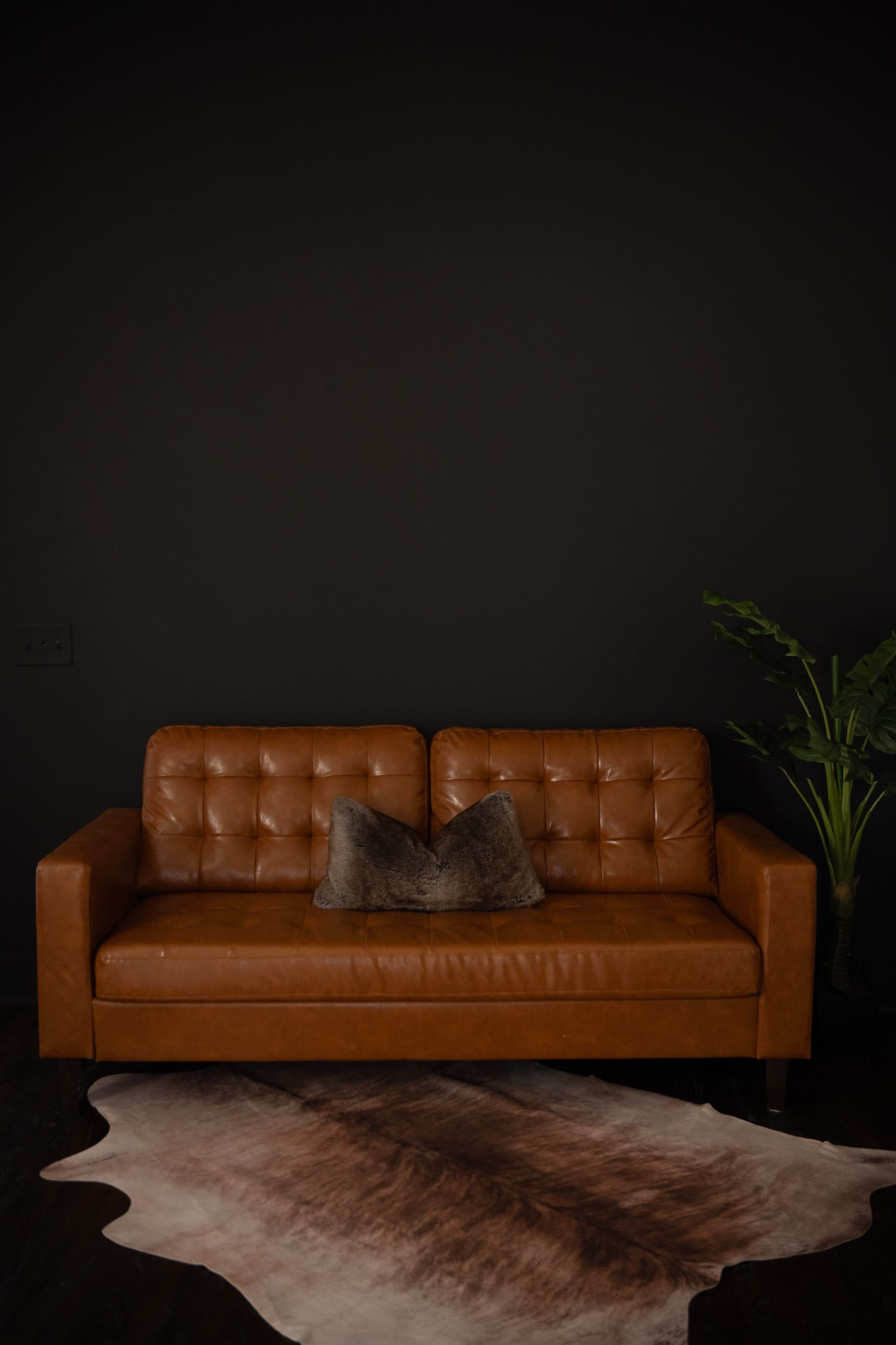 A brown leather sofa with tufted cushions is positioned against a dark wall, accompanied by a potted plant and a cowhide rug on the floor.