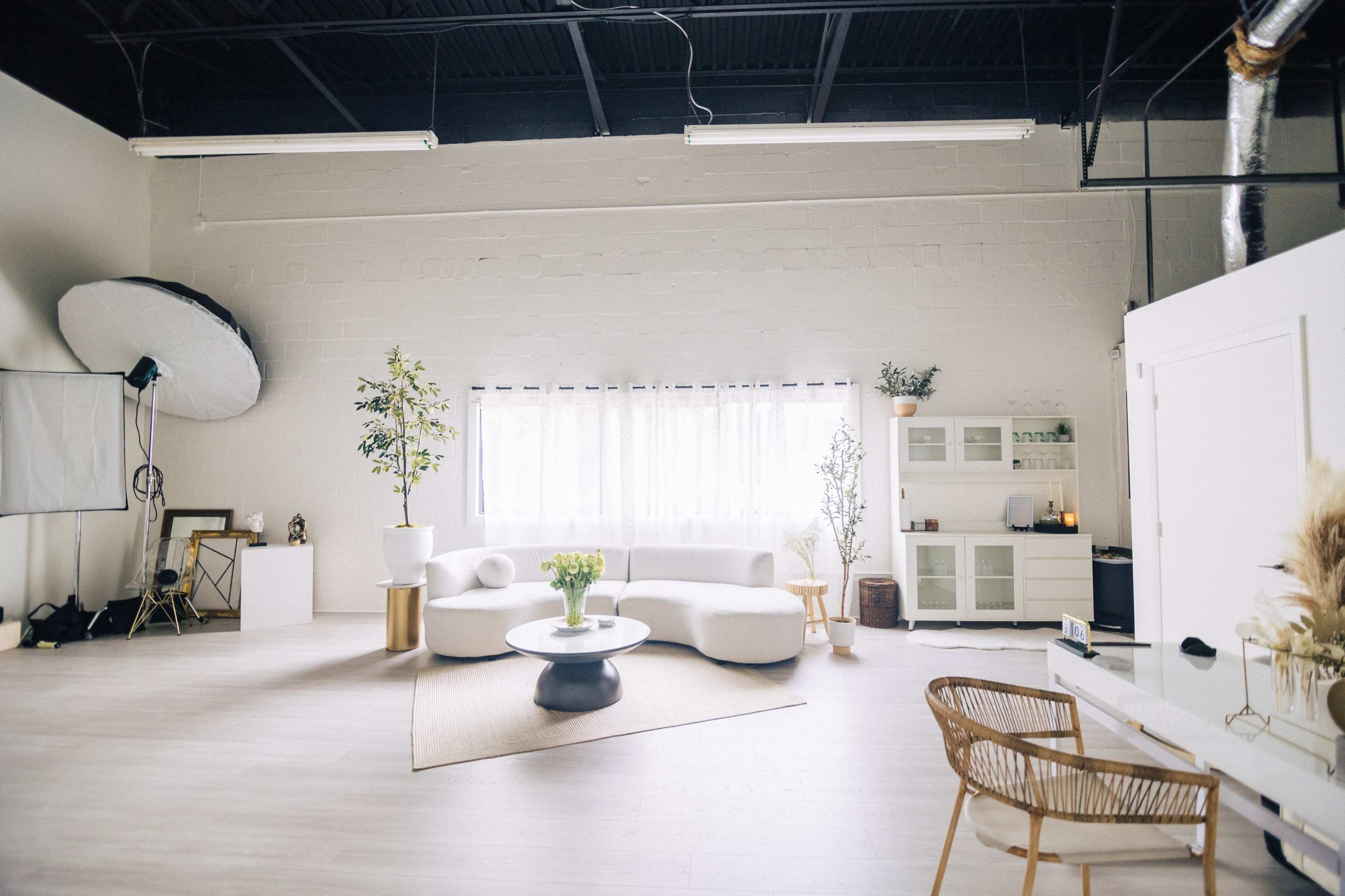 The image shows a spacious, well-lit studio with a white sofa, round coffee table, a large plant, and minimalist decor, including a shelf with various items.