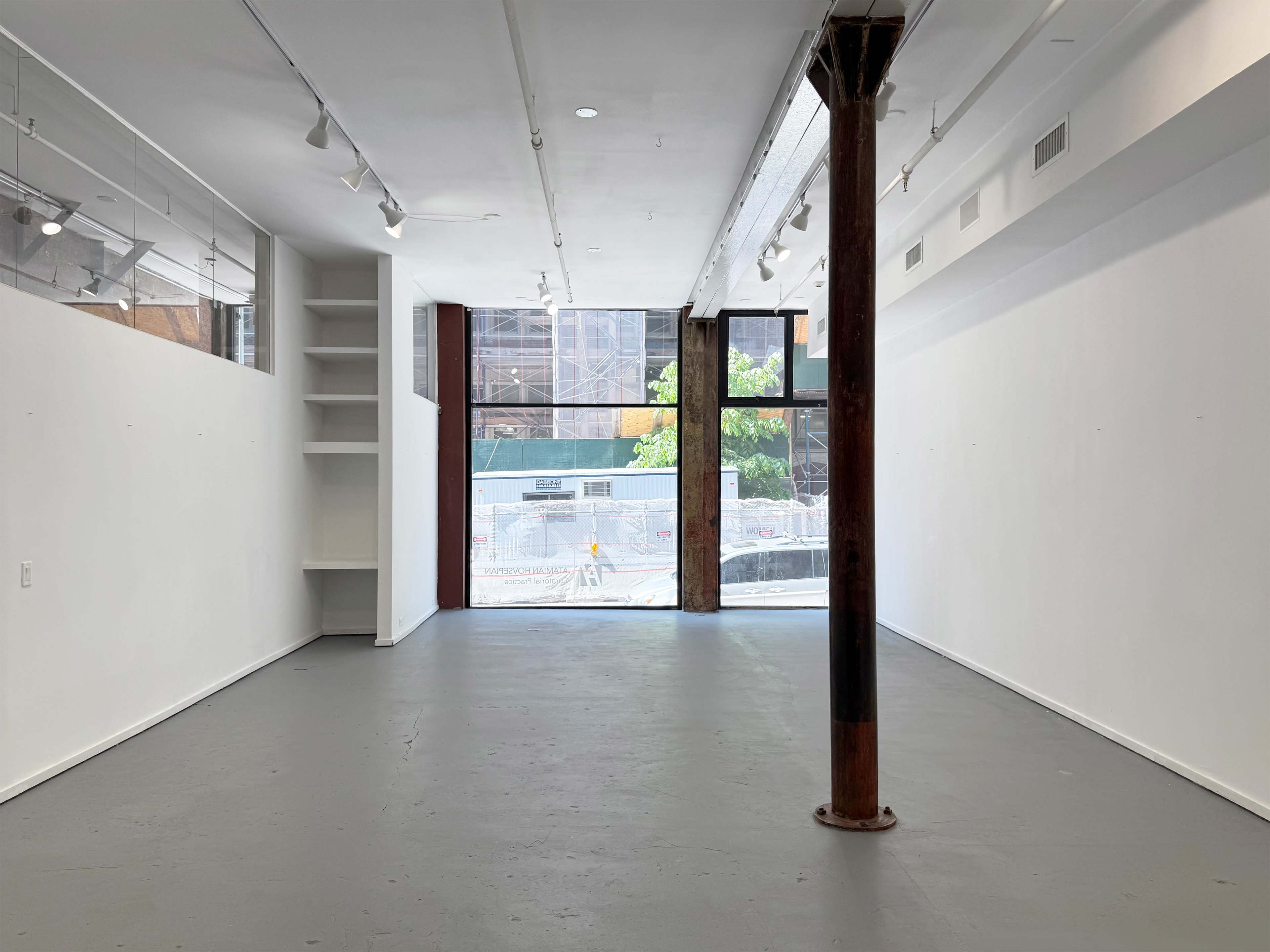 The image shows an empty interior space with white walls, a concrete floor, and a central support column, featuring large windows that overlook a construction view outside.