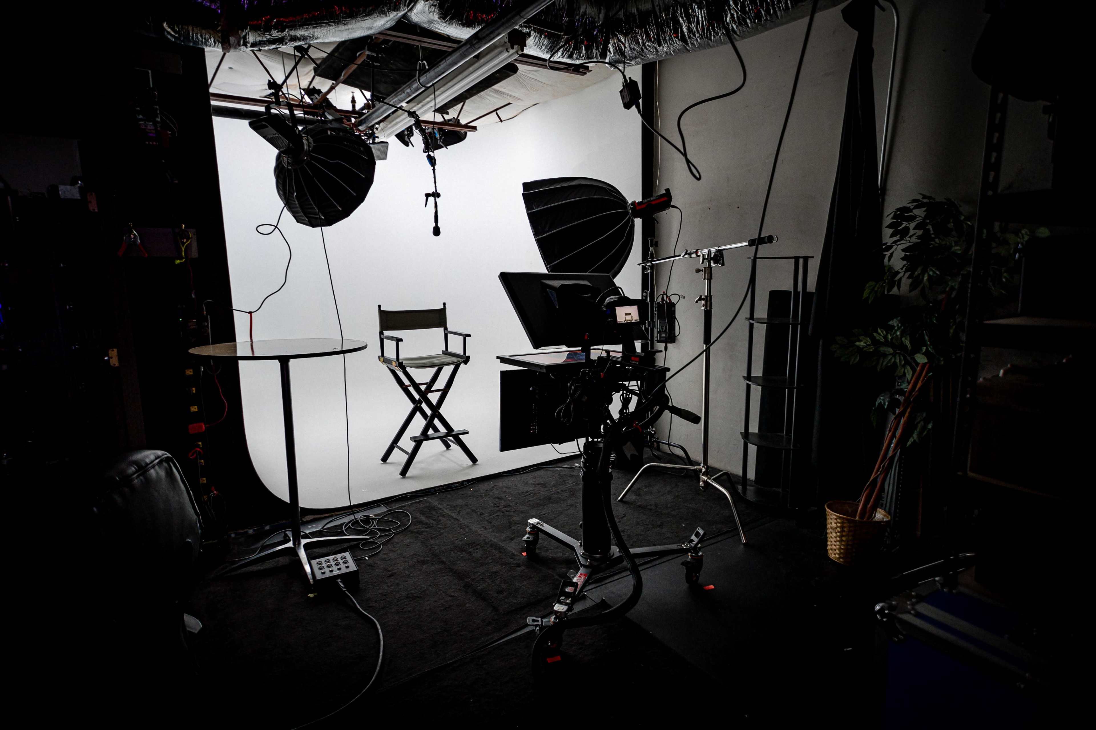 The image depicts a photography studio setup featuring a white background, a director's chair, lighting equipment, and a camera on a tripod.