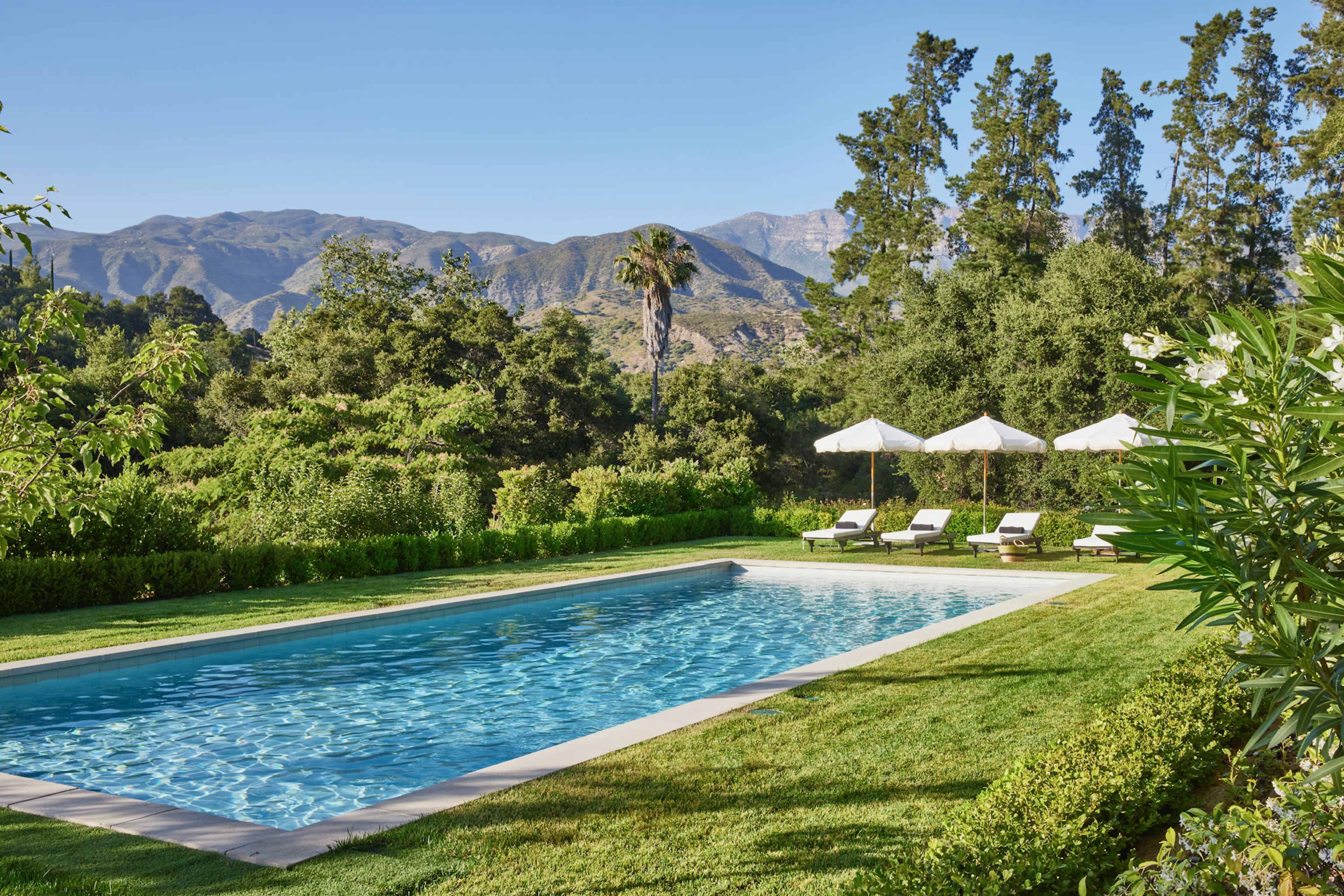 A rectangular swimming pool is surrounded by lush greenery and lounge chairs under white umbrellas, with mountains in the background.