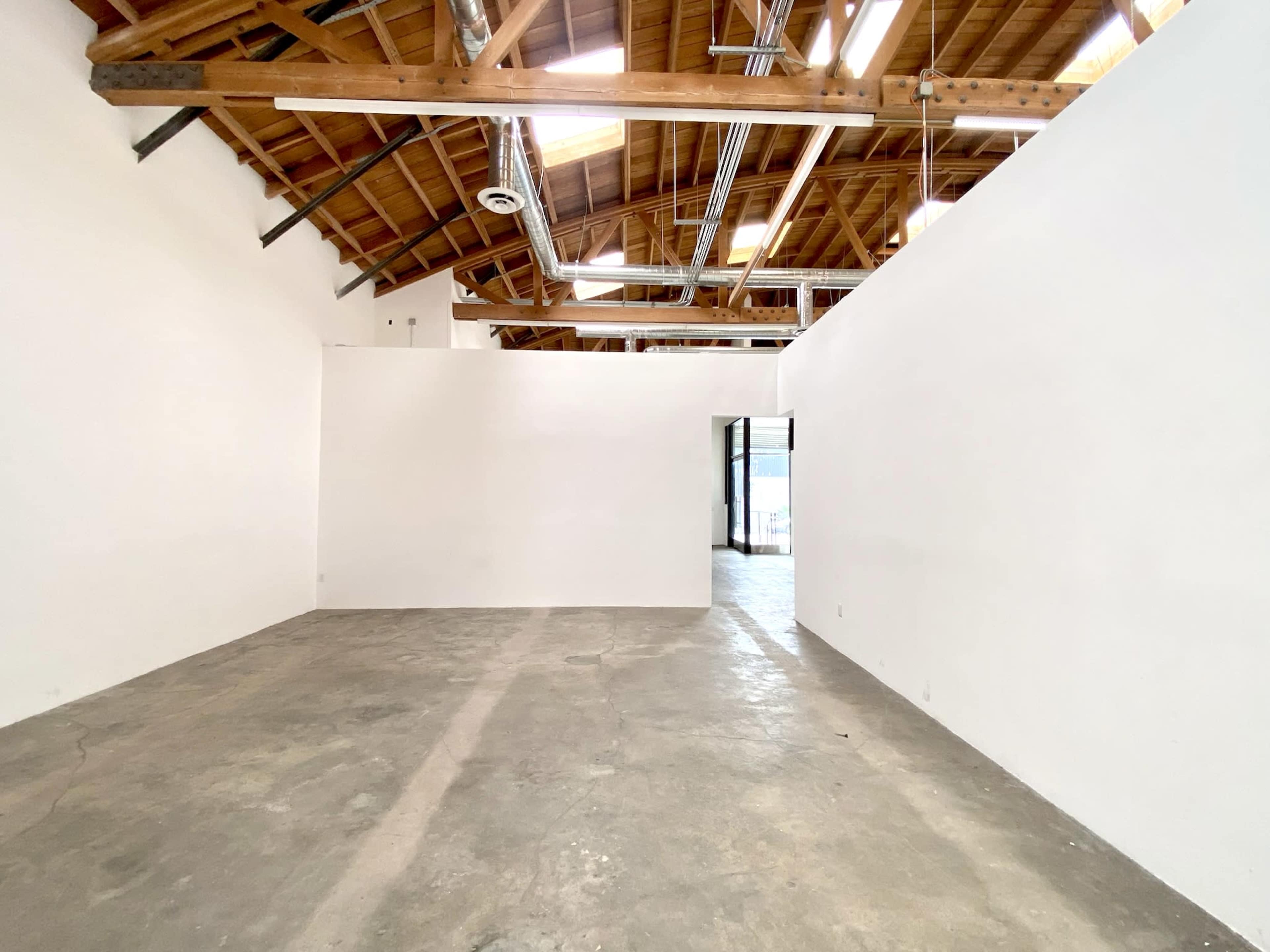 An empty room with white walls, a concrete floor, and exposed wooden beams in the ceiling.