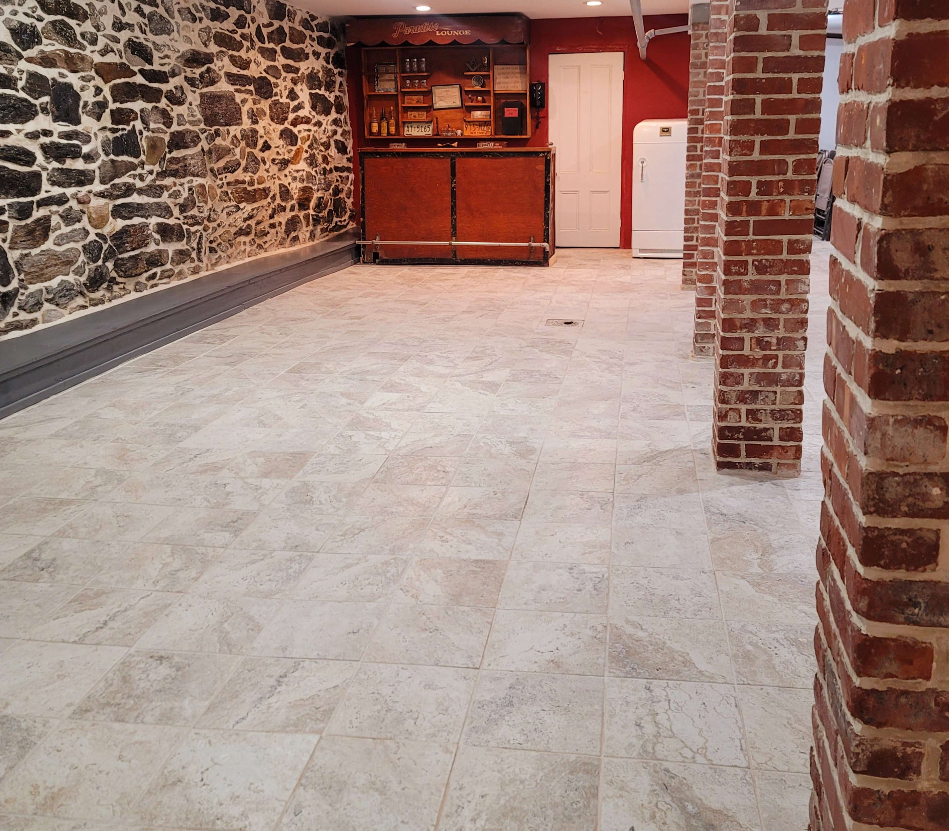 A spacious room with a tiled floor, stone walls on one side, and a wooden bar counter along with a refrigerator in the background.
