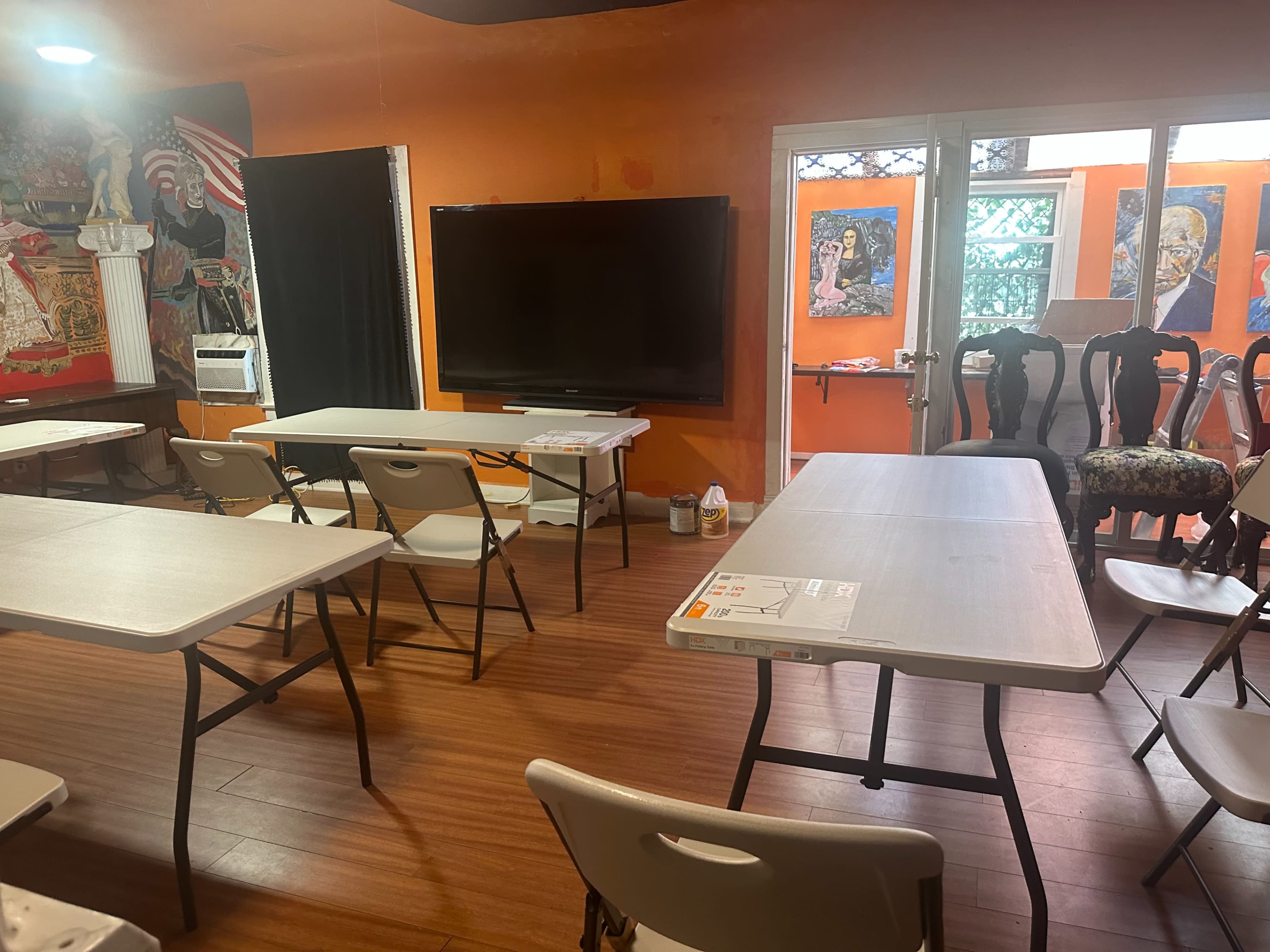The image shows a brightly colored room with several folding tables and chairs arranged for an event, featuring a large television on one wall and artwork on the others.