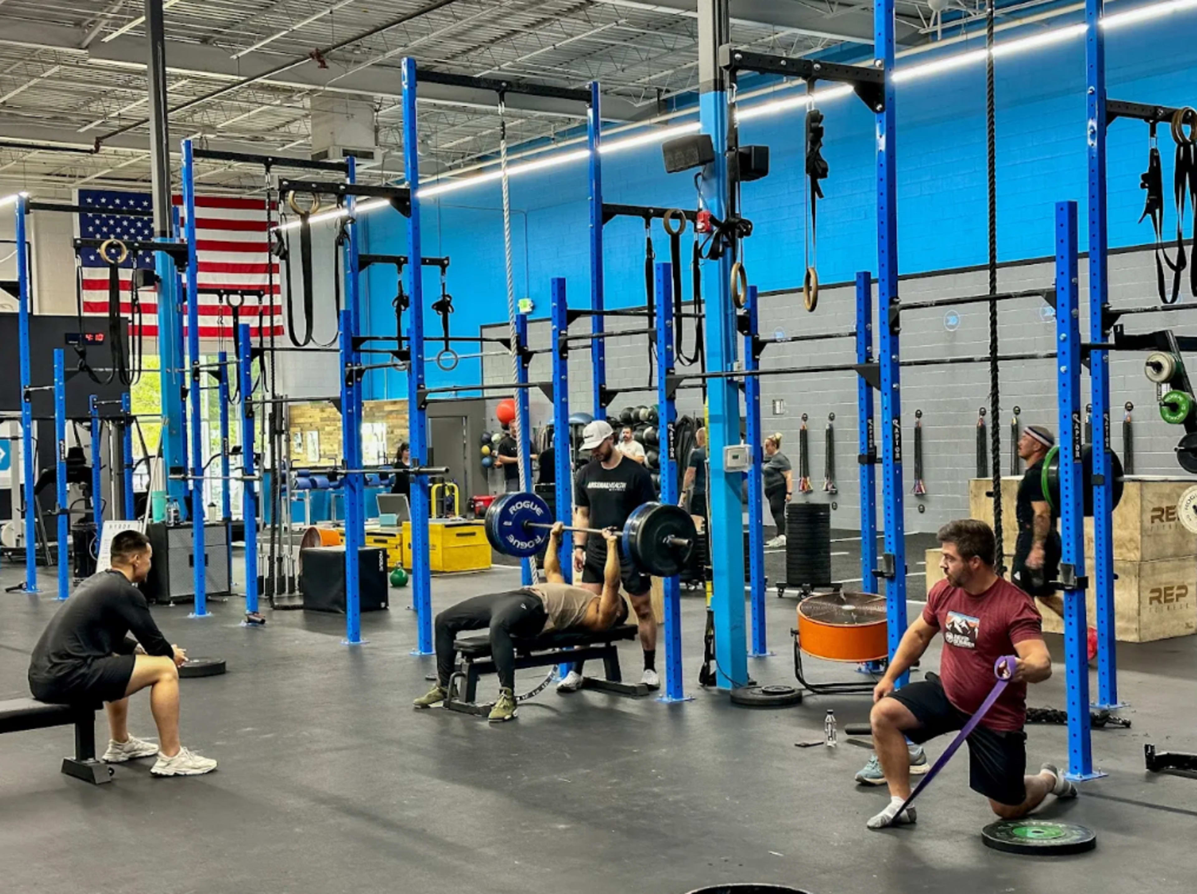 The image shows a busy gym with various individuals engaging in strength training and exercise activities among blue and black equipment.