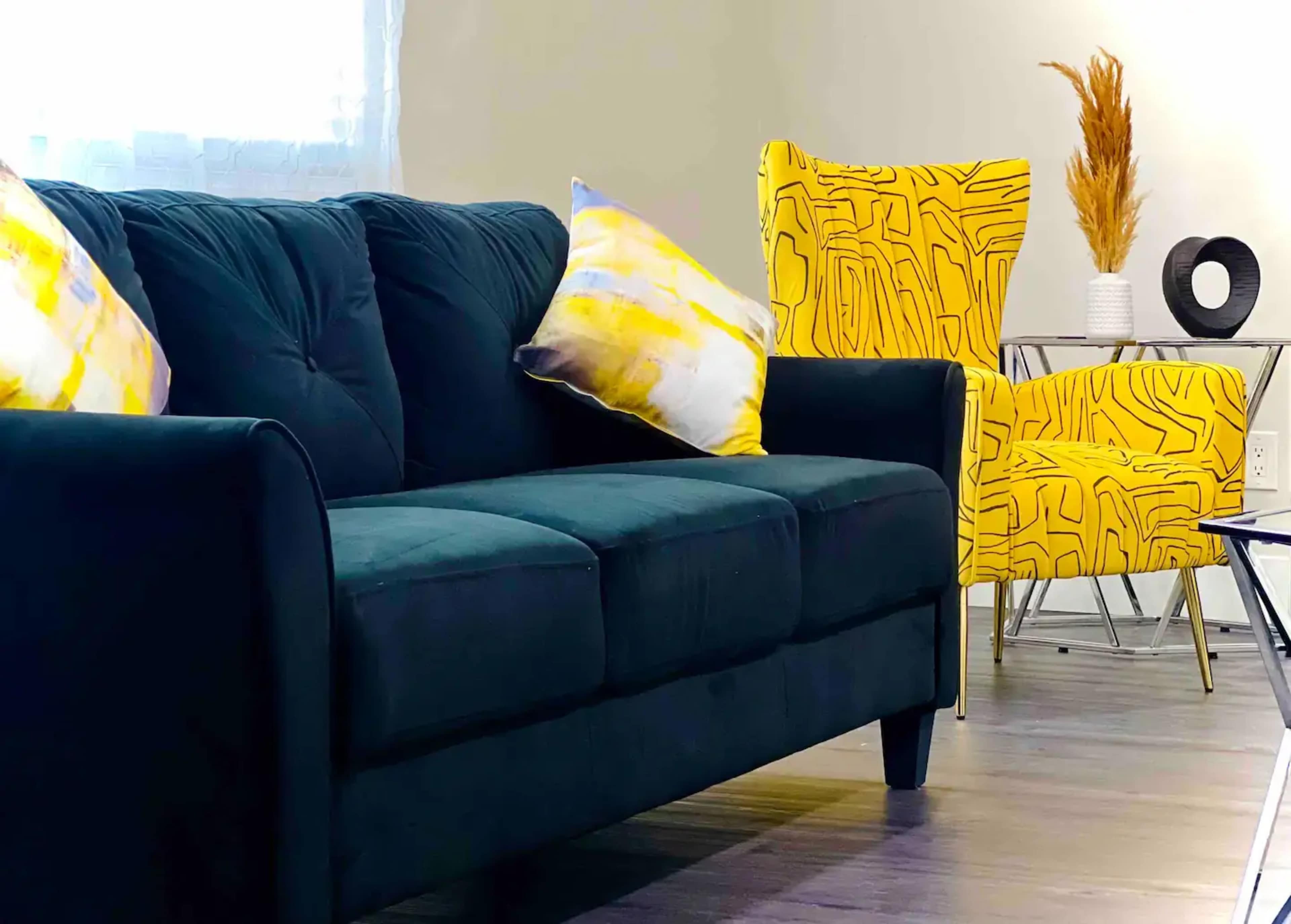 A dark green sofa with two decorative pillows and a yellow patterned accent chair nearby.