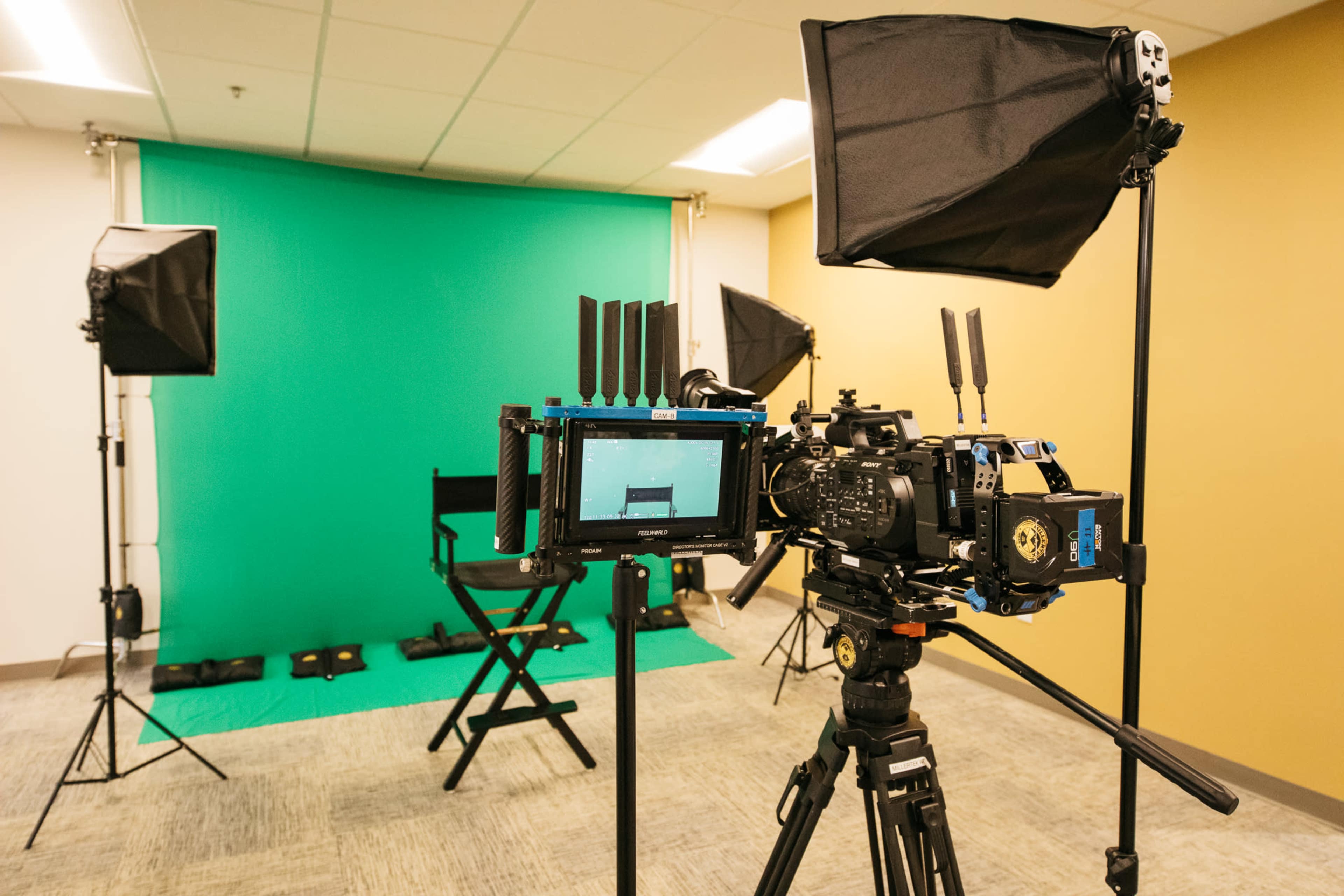 A video production setup featuring a camera on a tripod, a green backdrop, and studio lighting equipment positioned around a director's chair.