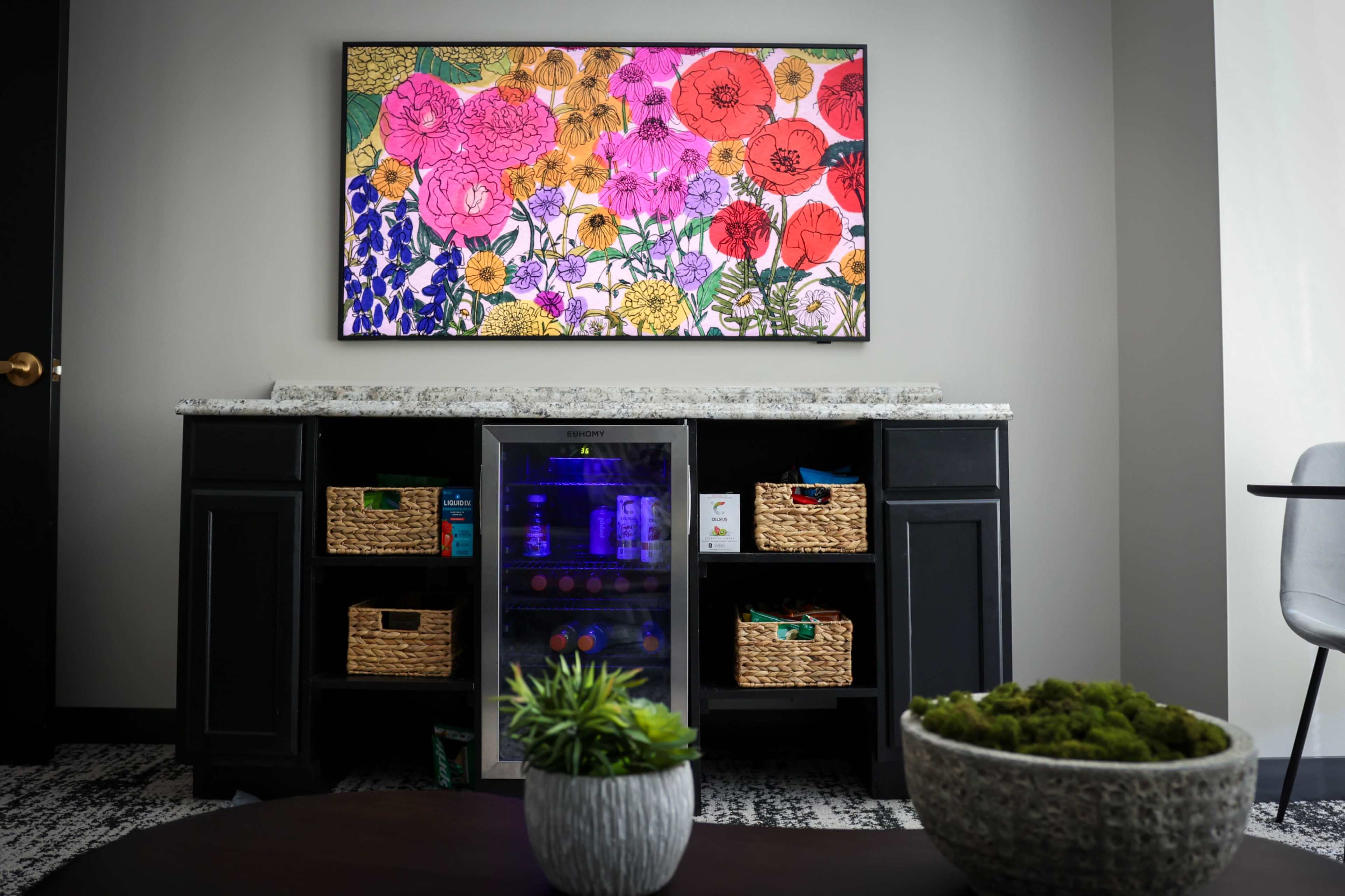 A modern seating area features a wall-mounted colorful floral artwork above a granite countertop with a mini fridge and woven basket storage.