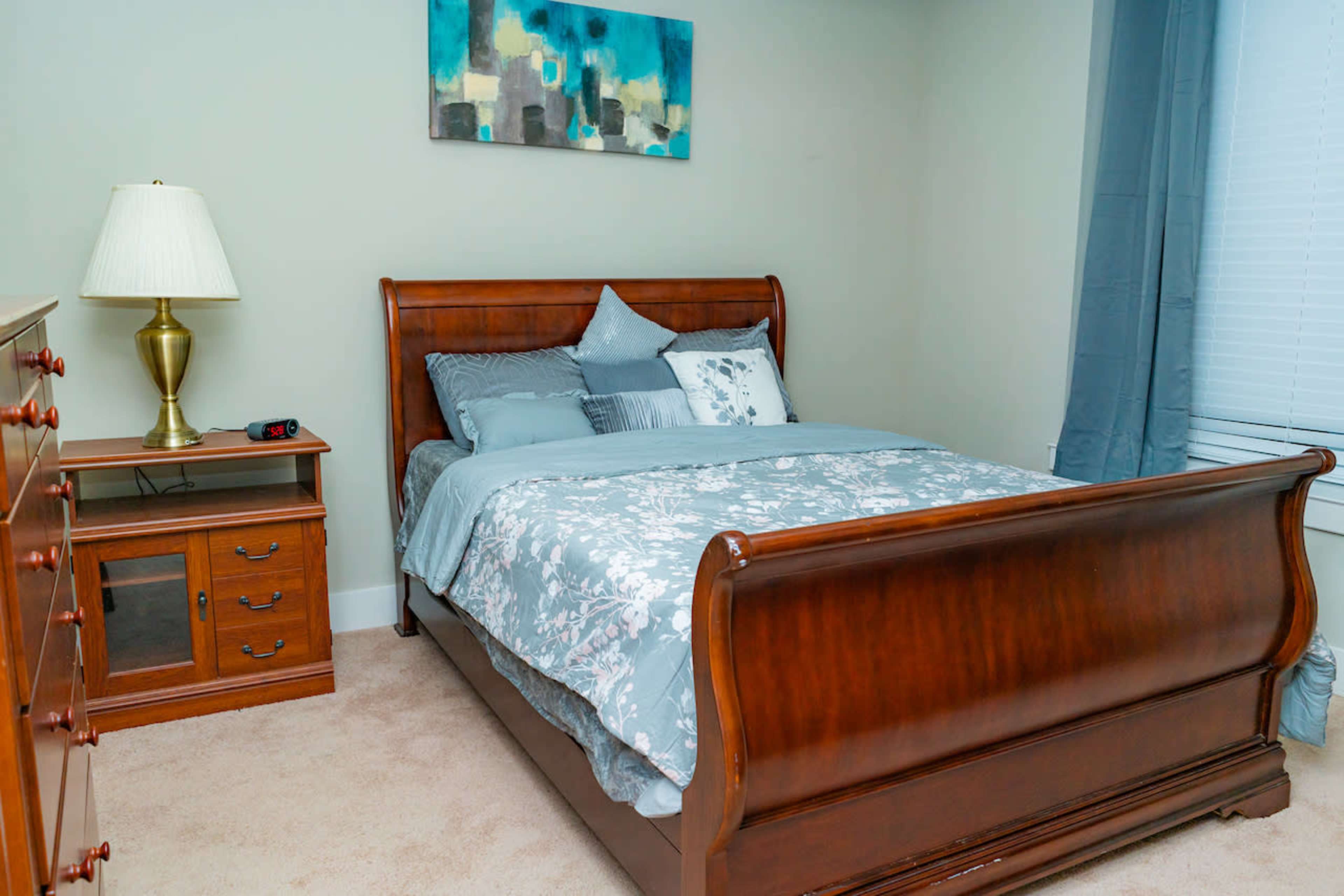 A neatly arranged bedroom features a wooden sleigh bed with a blue and gray patterned comforter, a nightstand with a lamp, and a dresser against the wall.