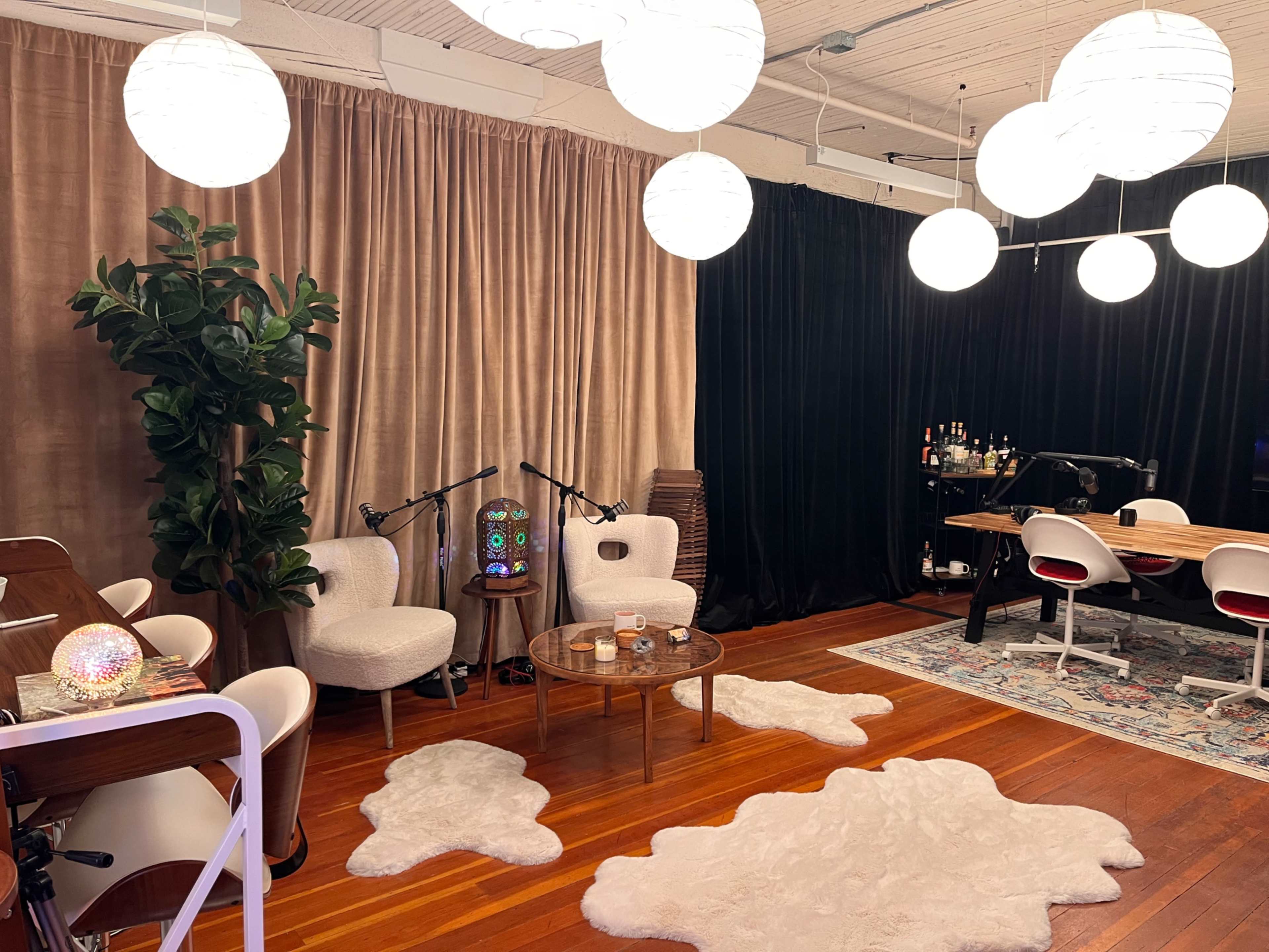 The image shows a modern interior space with a seating area featuring two white chairs, a round table, and a plant, alongside a recording setup with a bar area in the background.