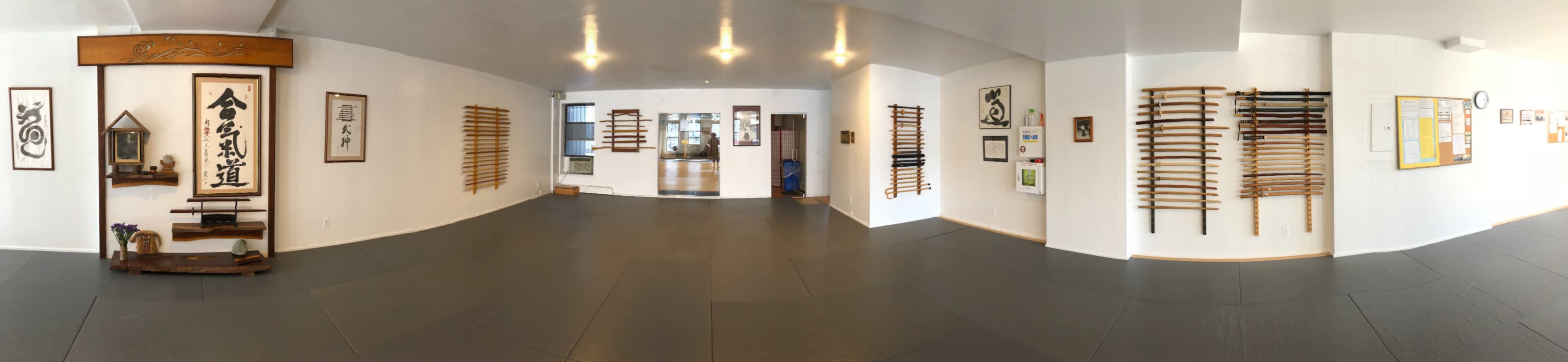 A panoramic view of a martial arts dojo featuring wooden training weapons displayed on the walls and an open, padded floor.