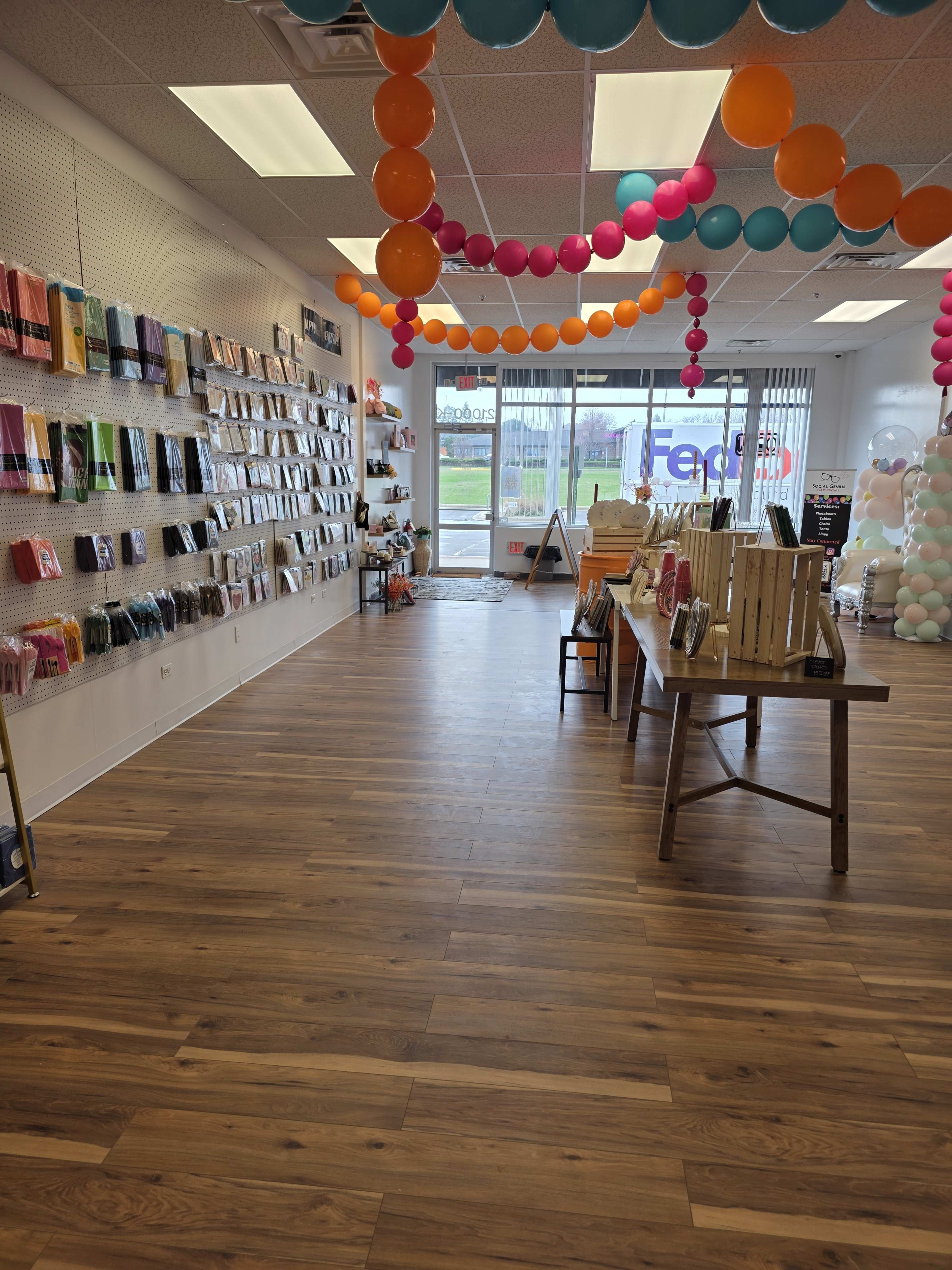 The image shows a brightly decorated retail space with colorful balloons, a display of products on the walls, and wooden tables arranged for merchandise in the center.
