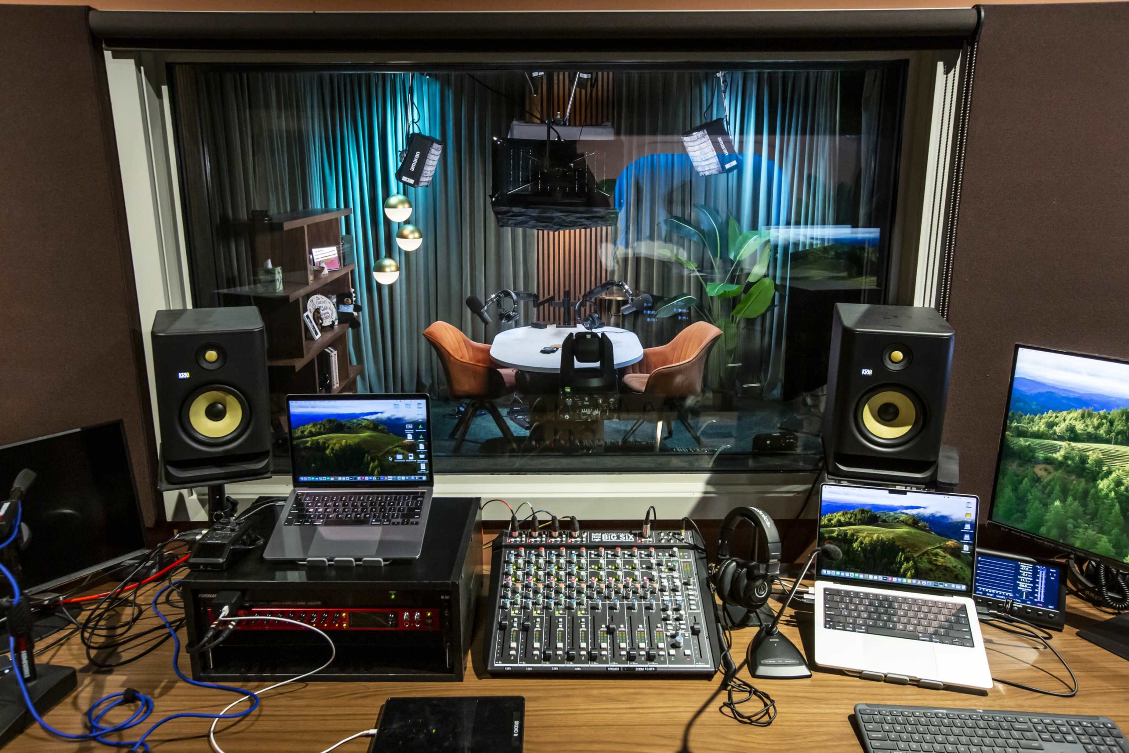 A recording studio features a control desk with audio equipment, laptops, and headphones, overlooking a brightly lit room set up for interviews or discussions.
