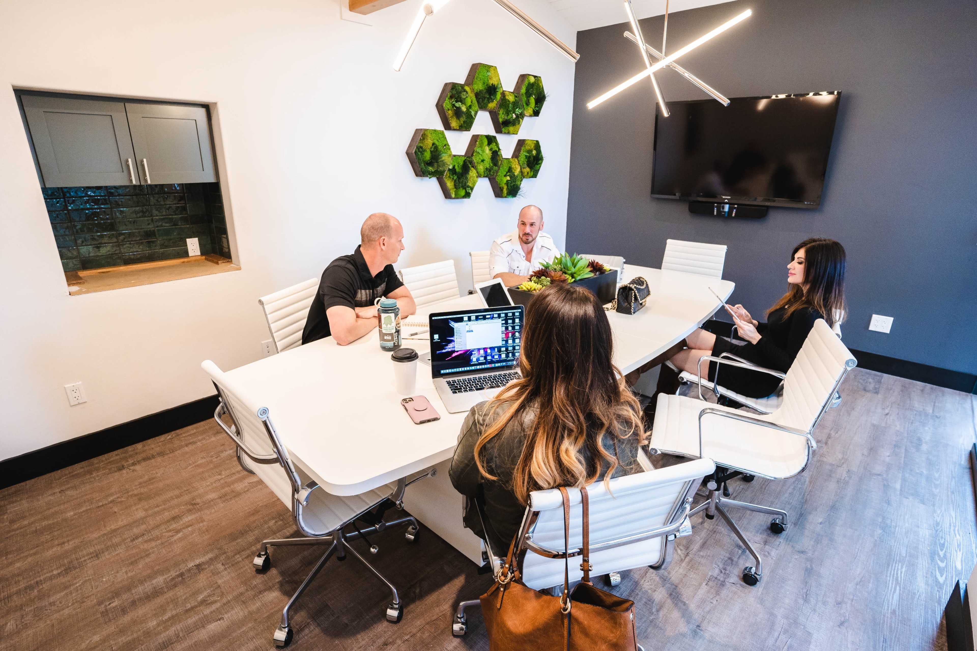 A group of five people are seated around a modern conference table in a well-lit meeting room, with a large screen and decorative wall elements in the background.