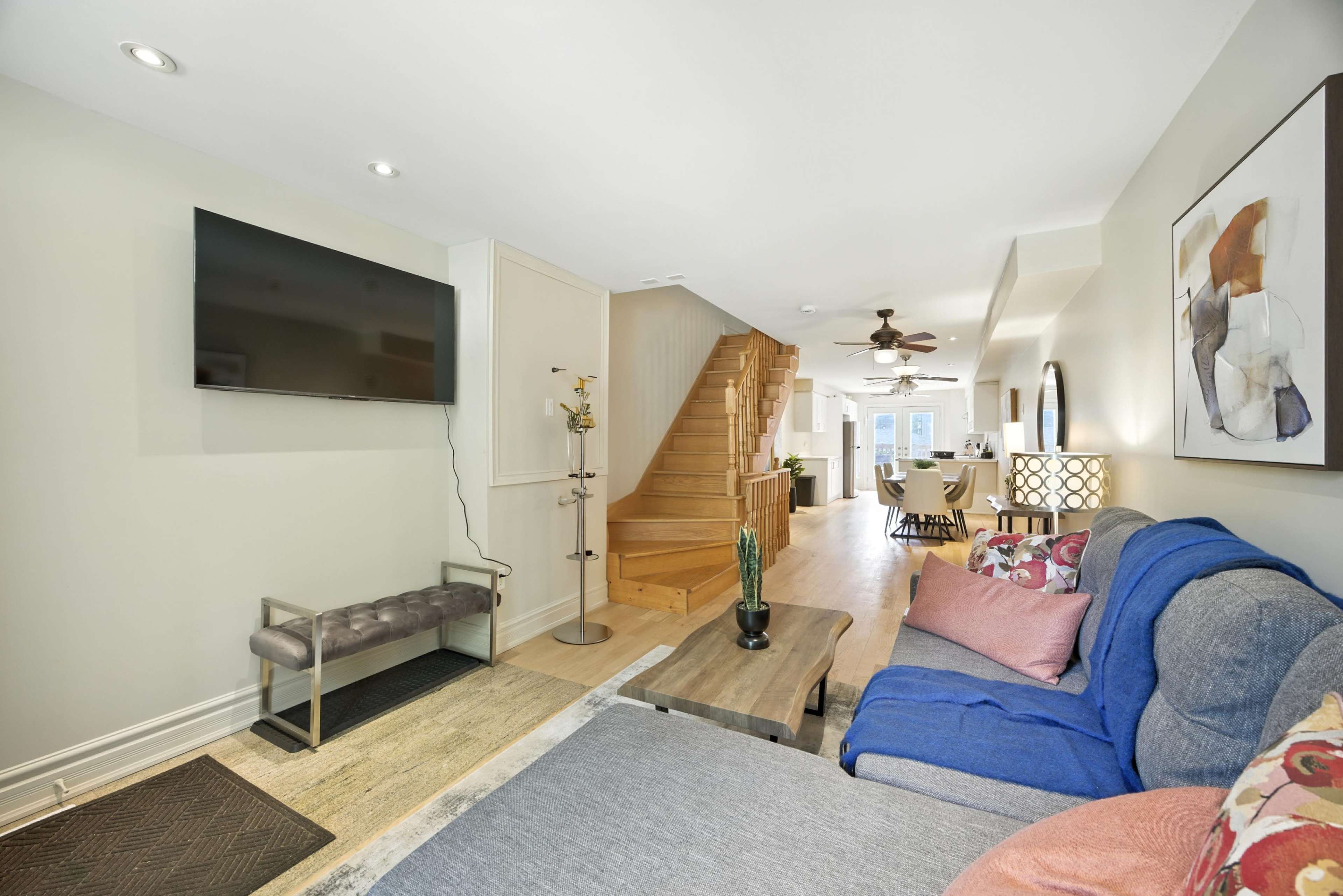 The image shows a modern living room with a sectional sofa, a coffee table, a wall-mounted television, and a staircase leading to the upper level.