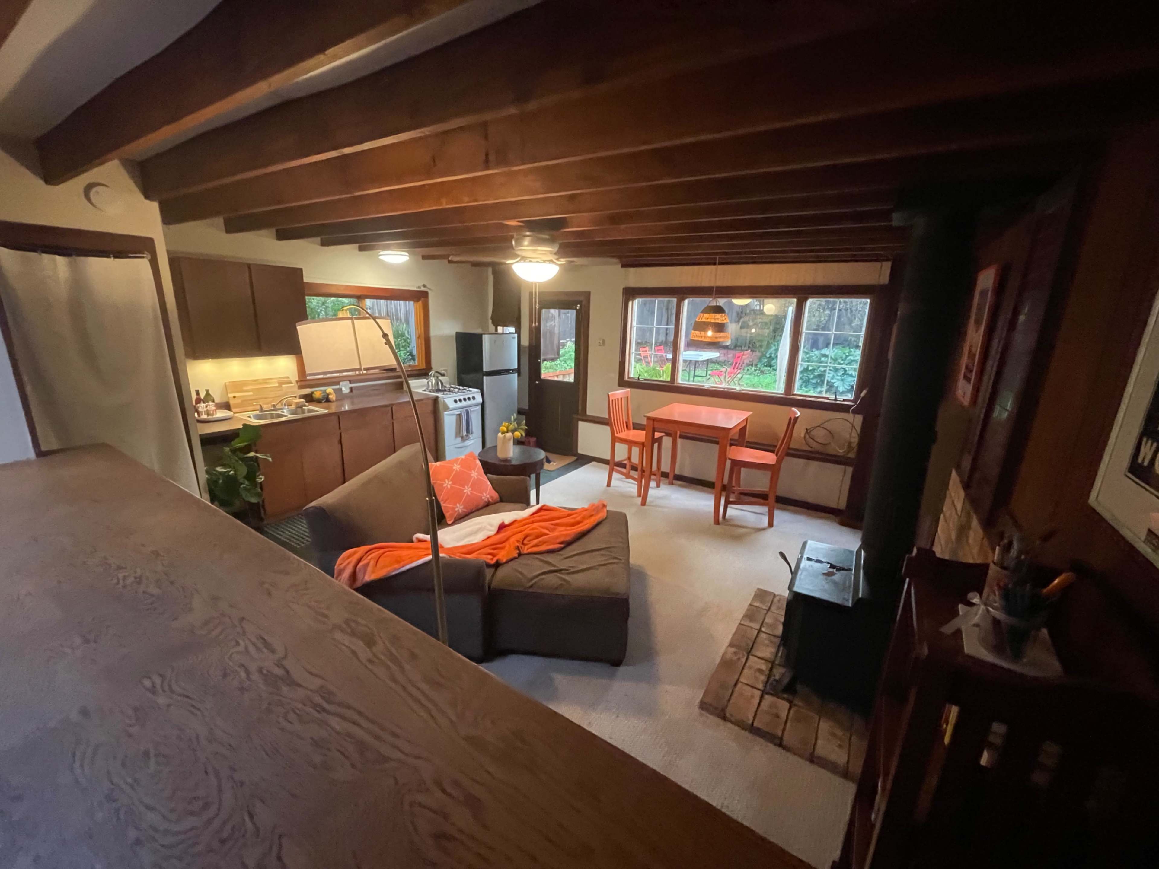 A cozy living space with a sectional couch, a dining table, and kitchen facilities, all featuring wooden beams and natural light from windows overlooking greenery.