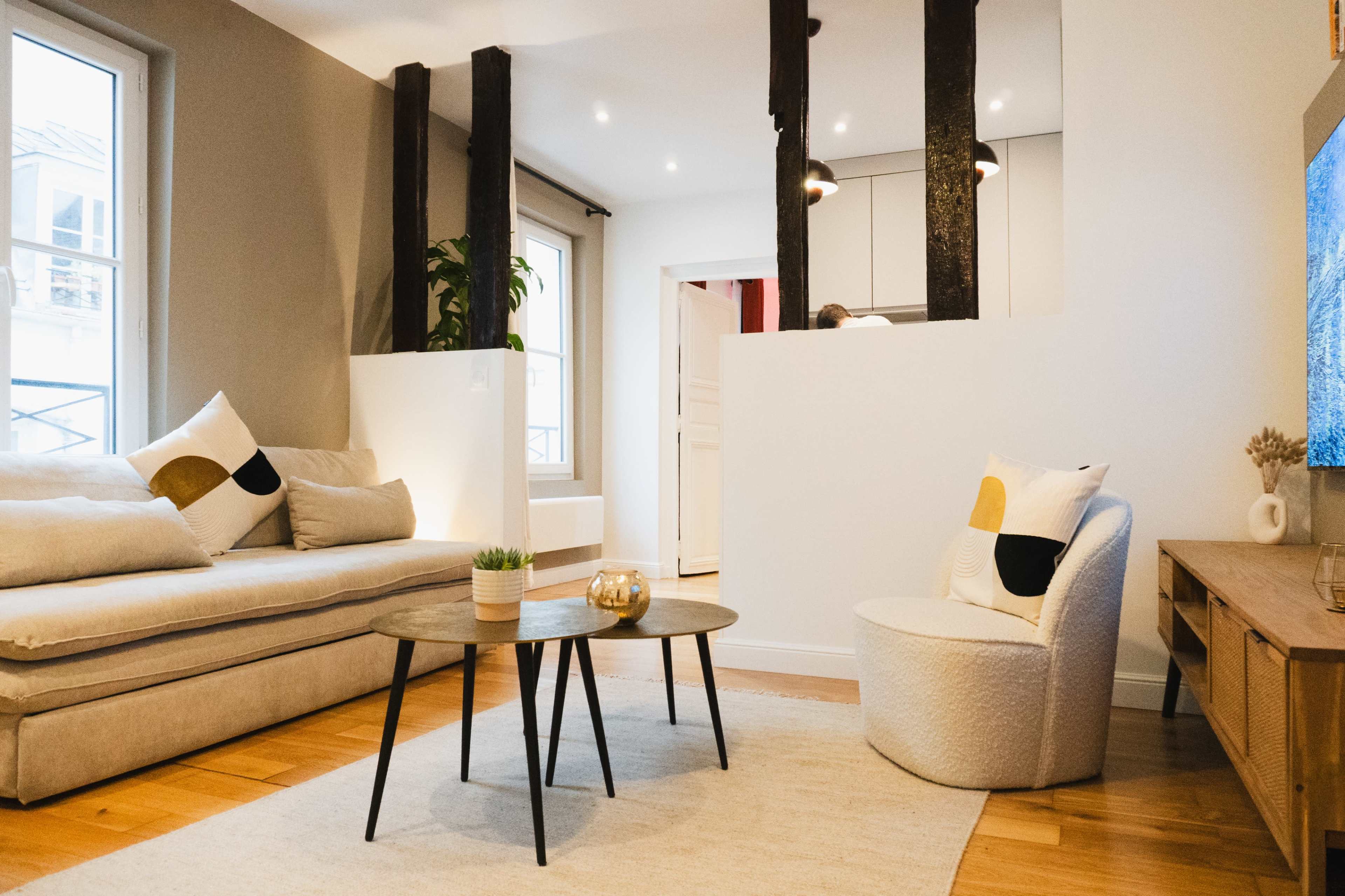 The image shows a modern living room featuring a beige sofa, a round coffee table, a white accent chair, and a television mounted on a wooden console.