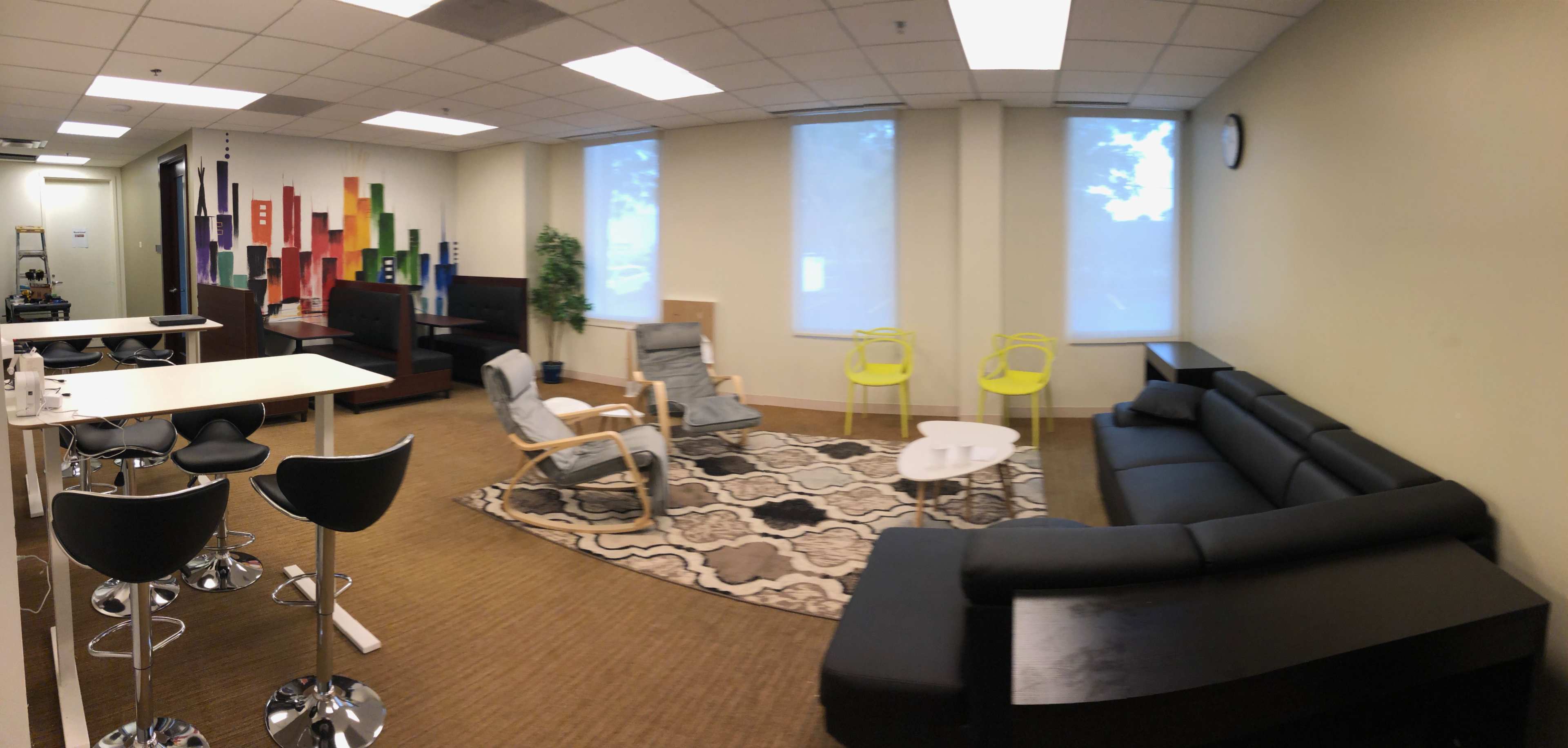 The image shows a spacious lounge area featuring a mix of seating arrangements, including black sofas, colorful chairs, and a round table on a patterned rug.