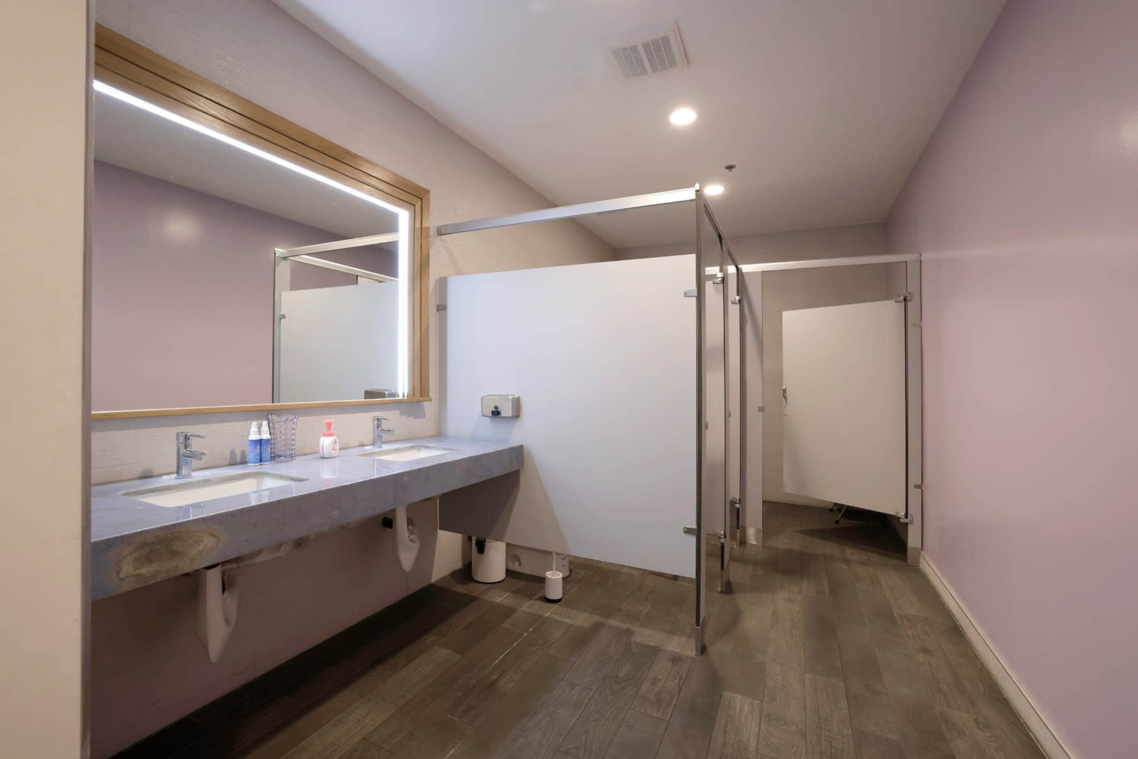 A women's restroom interior featuring two sinks, a large mirror, and three stalls with gray dividers.