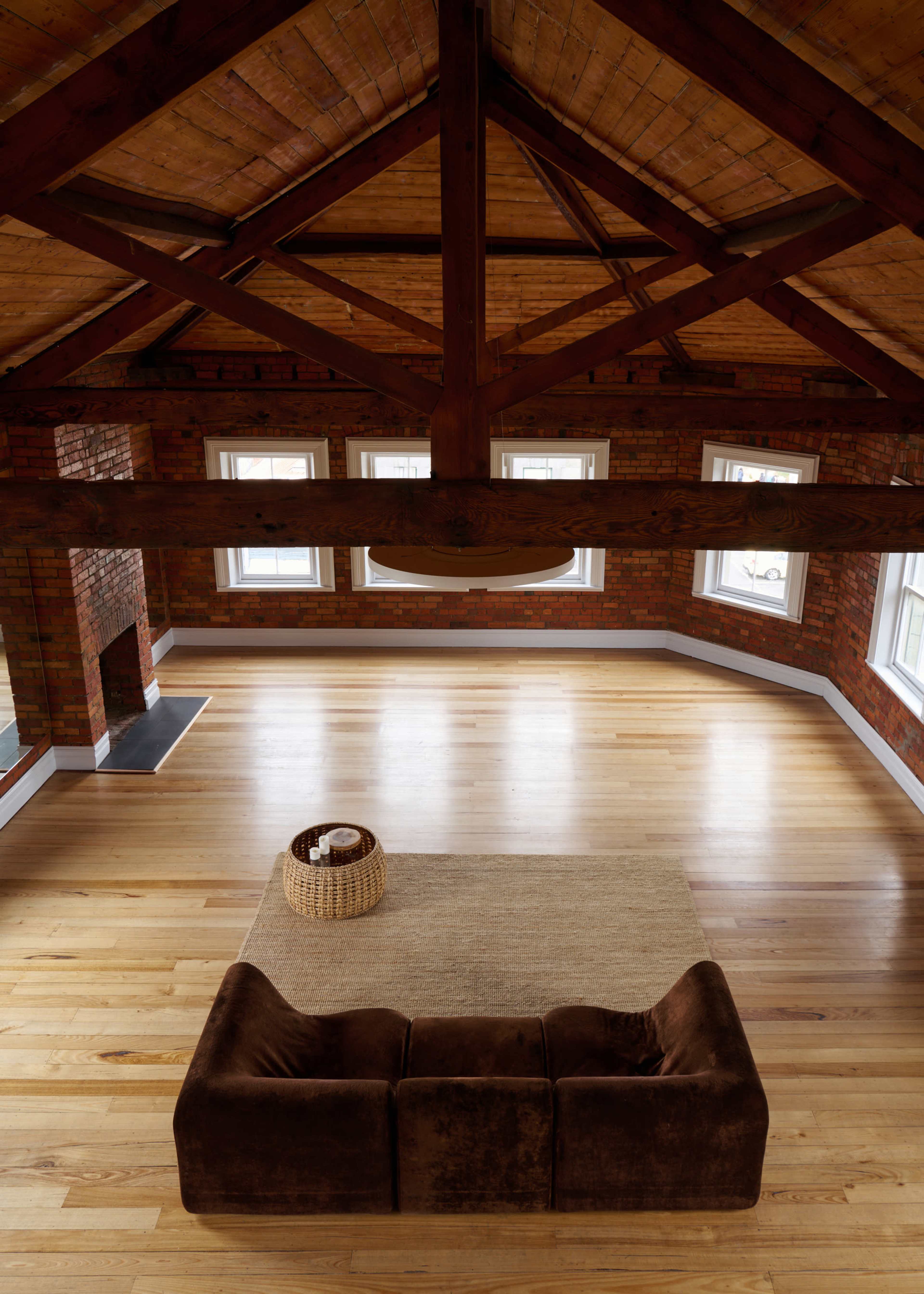 The image shows a spacious, modern living room with exposed wooden beams, large windows, and hardwood flooring, featuring a brown sofa and a woven basket on a rug.