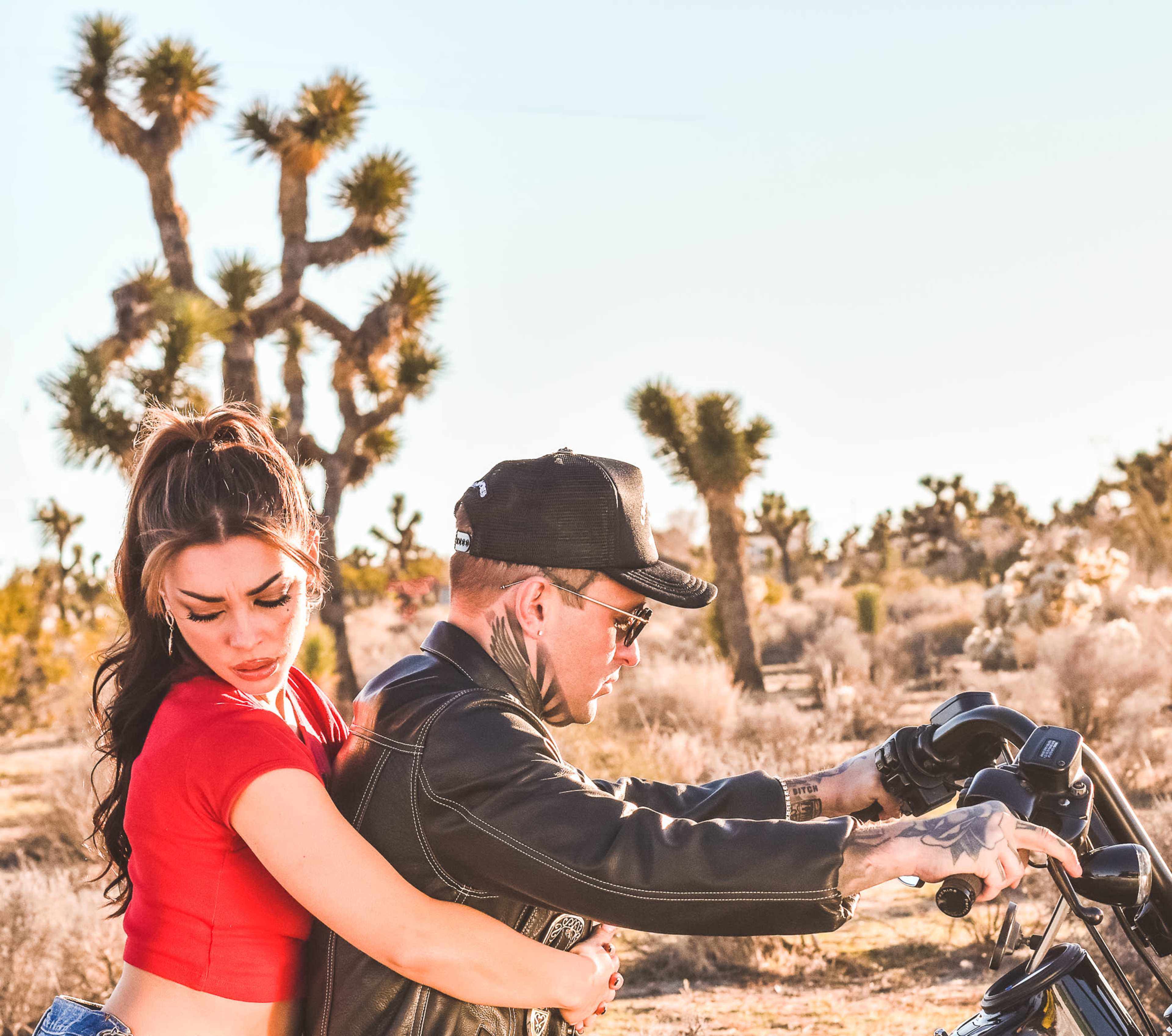 A woman in a red crop top embraces a man wearing a leather jacket and sunglasses as he sits on a motorcycle in a desert landscape with Joshua trees.