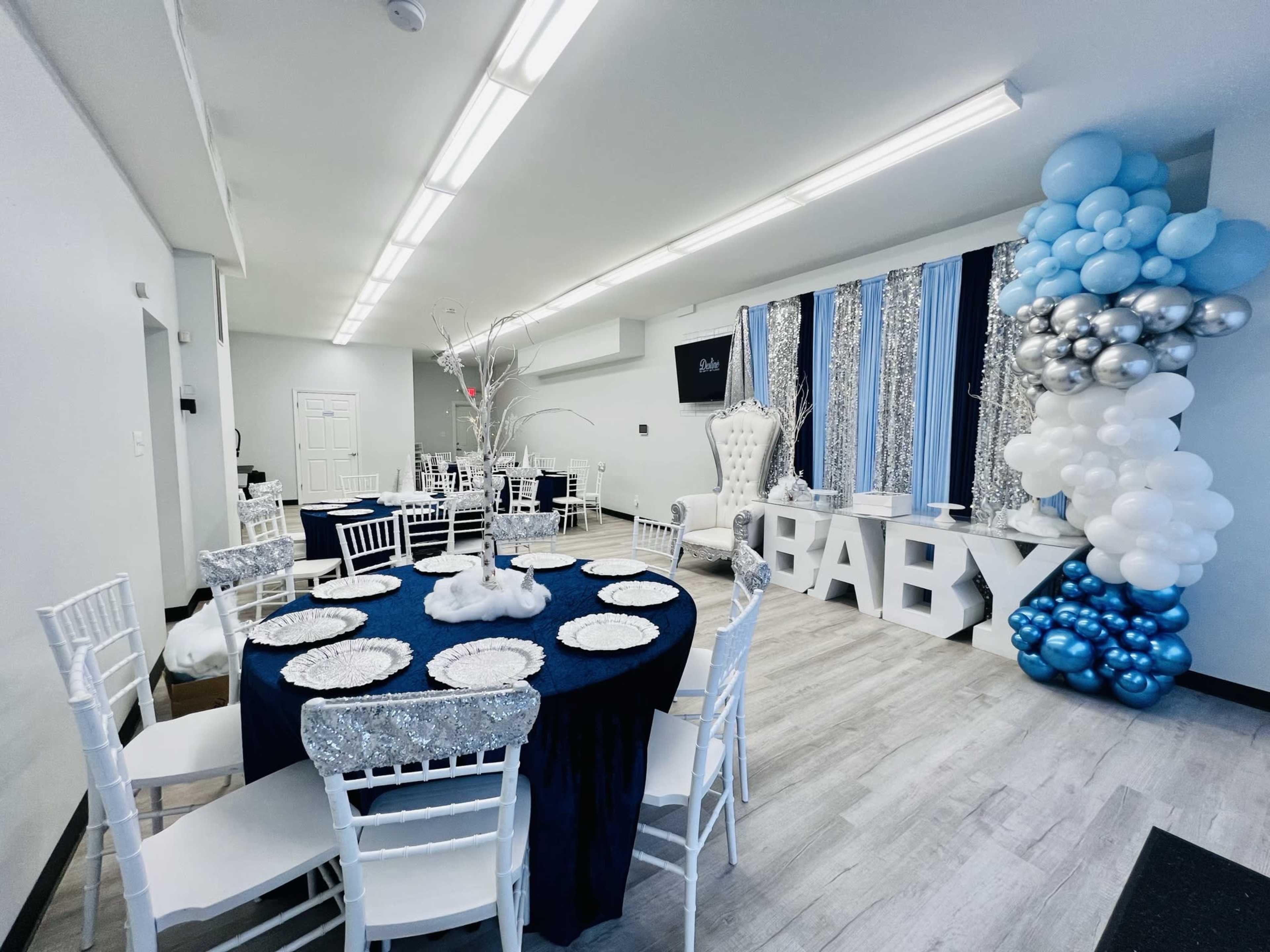The image shows a decorated event space with white chairs around a navy blue tablecloth, featuring a large "BABY" sign and a balloon arrangement in shades of blue and silver.